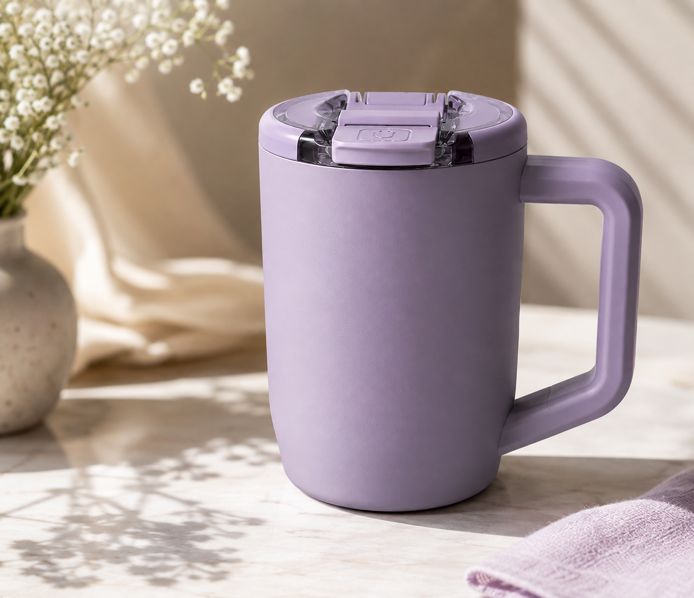 A lavender insulated travel mug with a flip-top lid on a white marble surface, next to a vase with white flowers and a folded pink cloth.