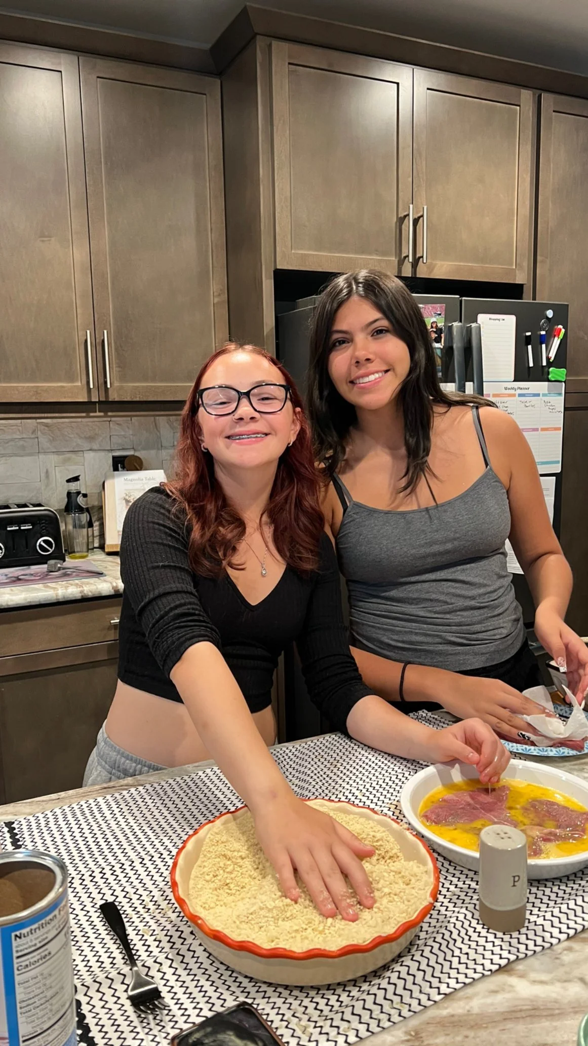 Two women in a kitchen preparing food together. One woman is pouring bread crumbs on meat in a baking dish, and the other is smiling at the camera. There are various kitchen items and a menu board on the fridge behind them.