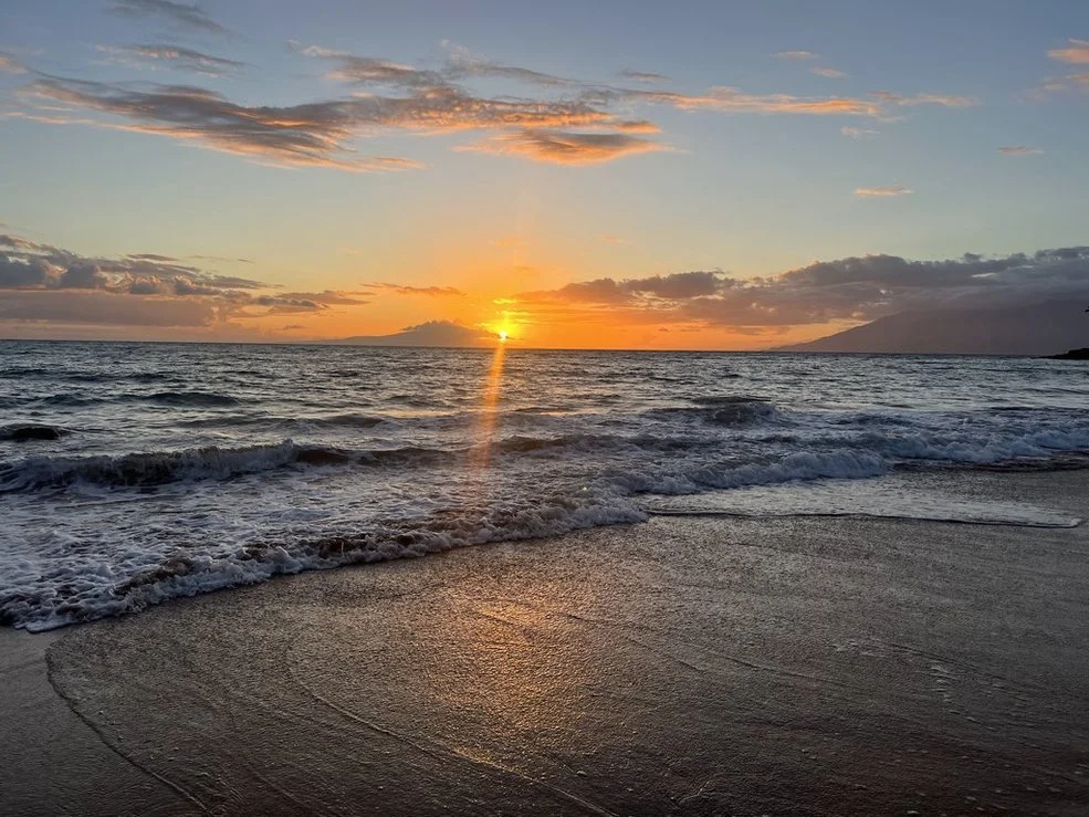 Sunset over the ocean with waves crashing on a sandy beach, partly cloudy sky, and distant landmass.