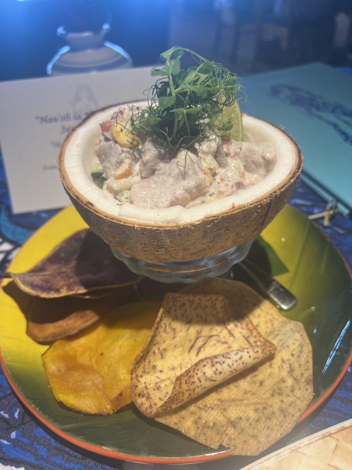 A tropical dish served in a coconut shell garnished with herbs and lime, accompanied by fried plantains and tortillas on a colorful plate.