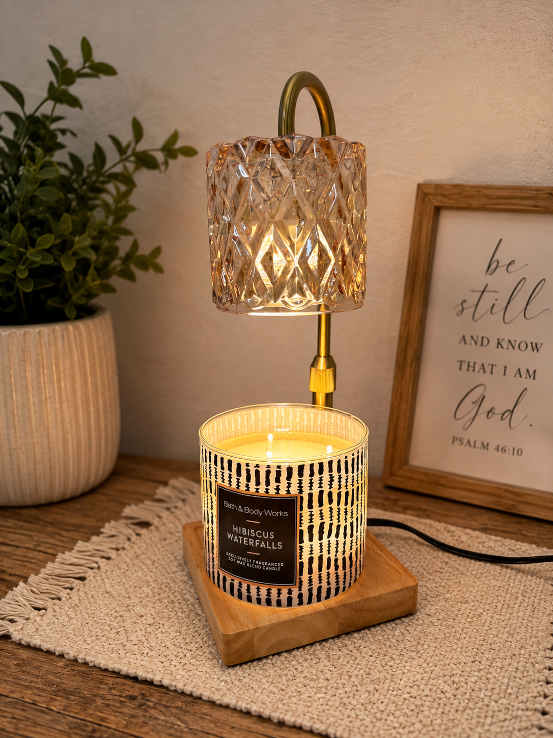 A decorative table lamp with a textured glass shade and a brass stem, placed on a wooden tray with a lit candle inside. A framed quote and a potted plant are in the background on a wooden surface.