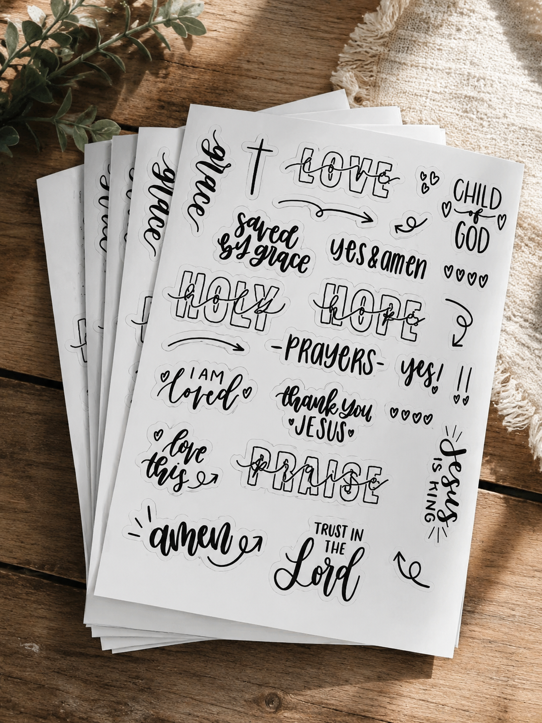 A sheet of black and white devotional stickers with religious phrases and words like 'Love,' 'Hope,' 'Prayers,' 'Thank you Jesus,' 'Child of God,' and various decorative elements, placed on a wooden surface with a plant and fabric nearby.