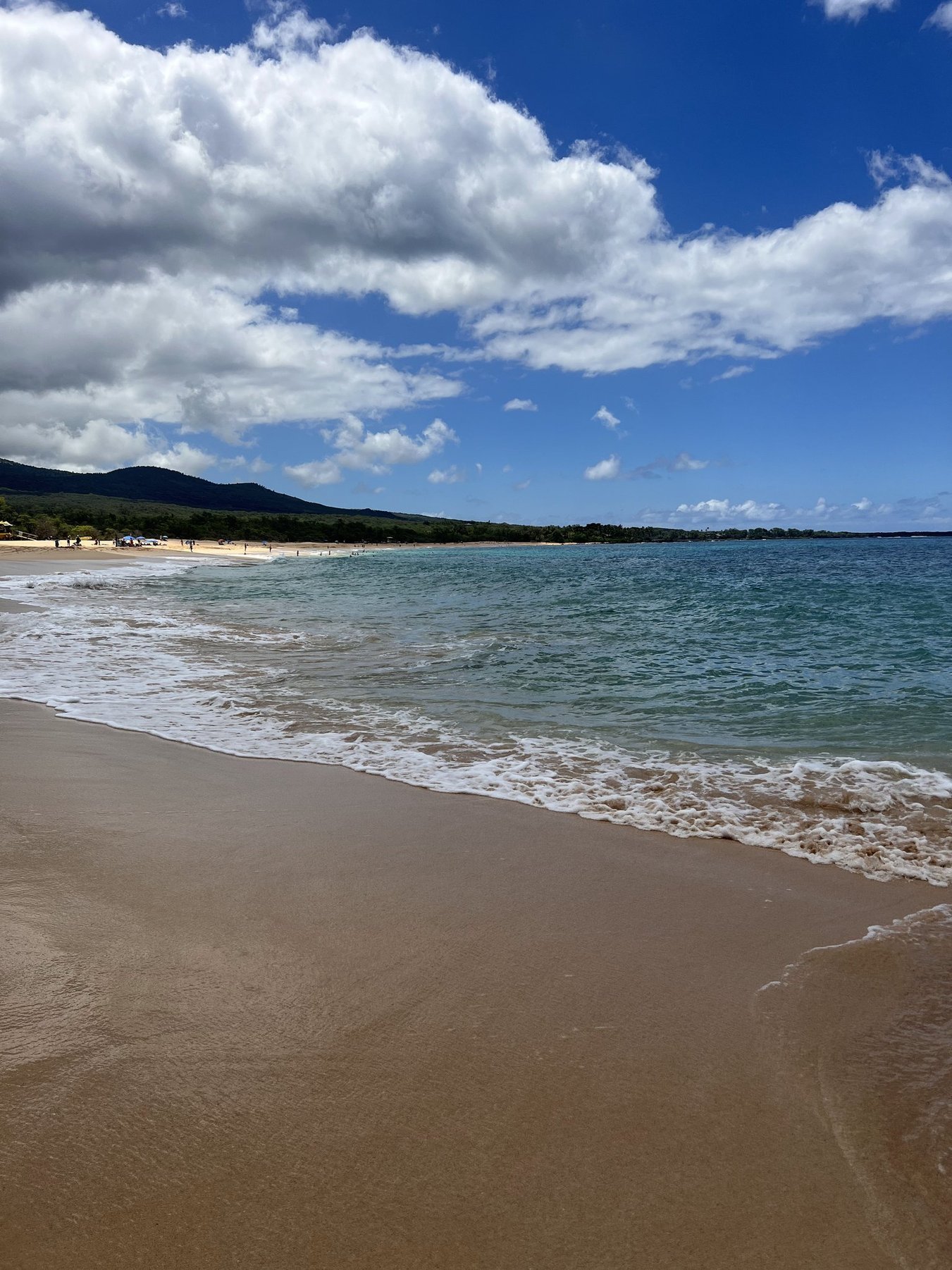 A sandy beach with gentle waves, people in the distance, green hills, and partly cloudy sky.