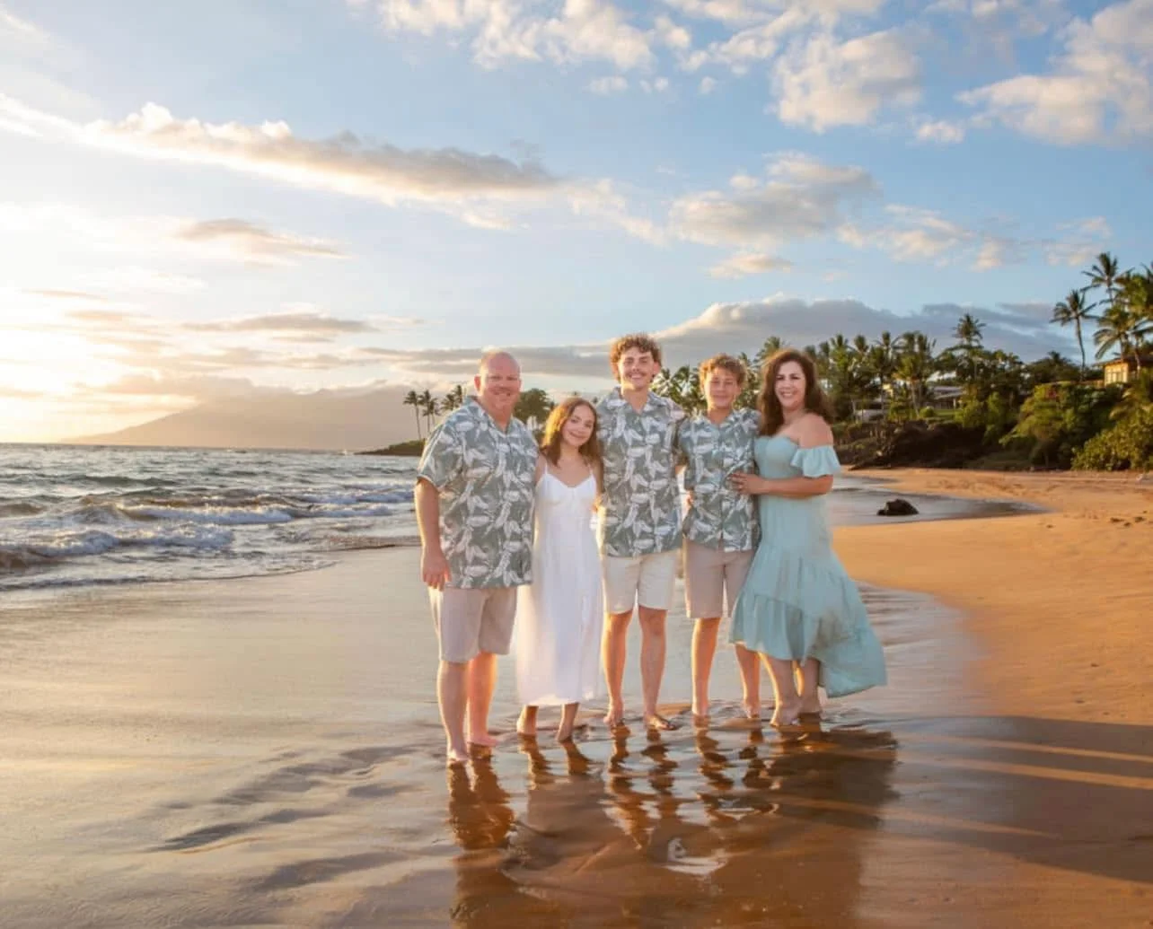 A family of six standing on a beach at sunset, wearing casual tropical clothing, with palm trees and a mountain in the background.
