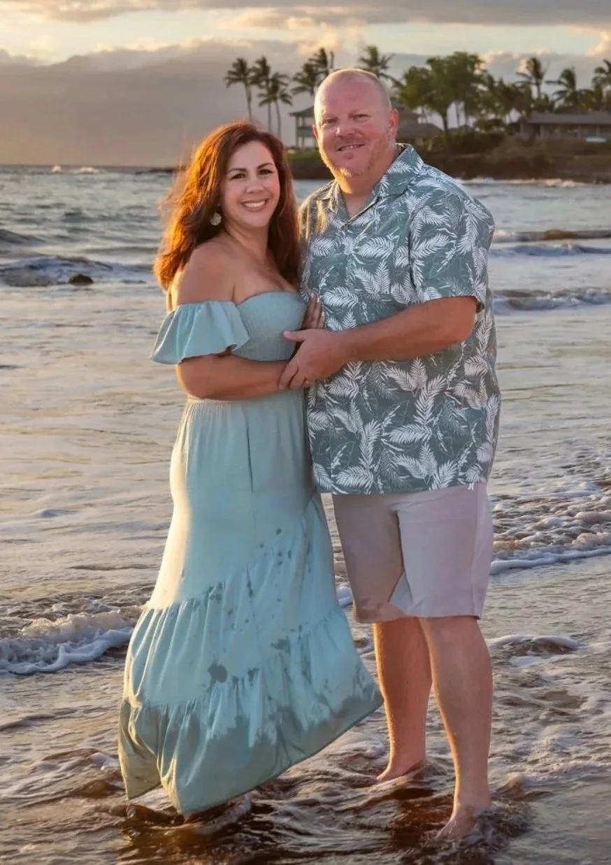 A couple standing barefoot in the shallow ocean water at sunset, smiling and embracing, with palm trees and houses in the background.