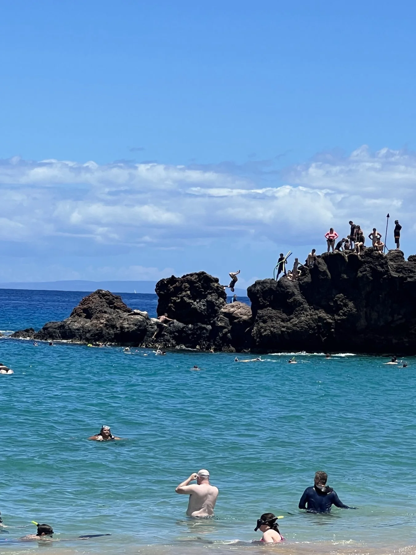 People enjoying a day at the beach with rocks and the ocean, some standing on rocks, swimming, and sunbathing under a partly cloudy sky.