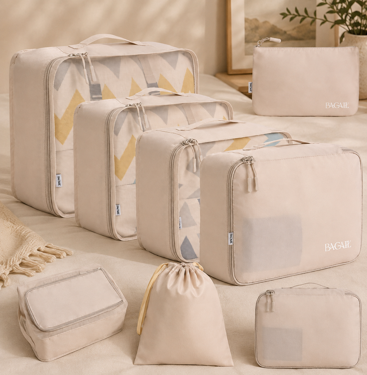 Multiple beige and patterned packing cubes and bags, including large, medium, small sizes, a zippered pouch, and a drawstring bag, arranged on a beige bed with a framed picture and a potted plant in the background.