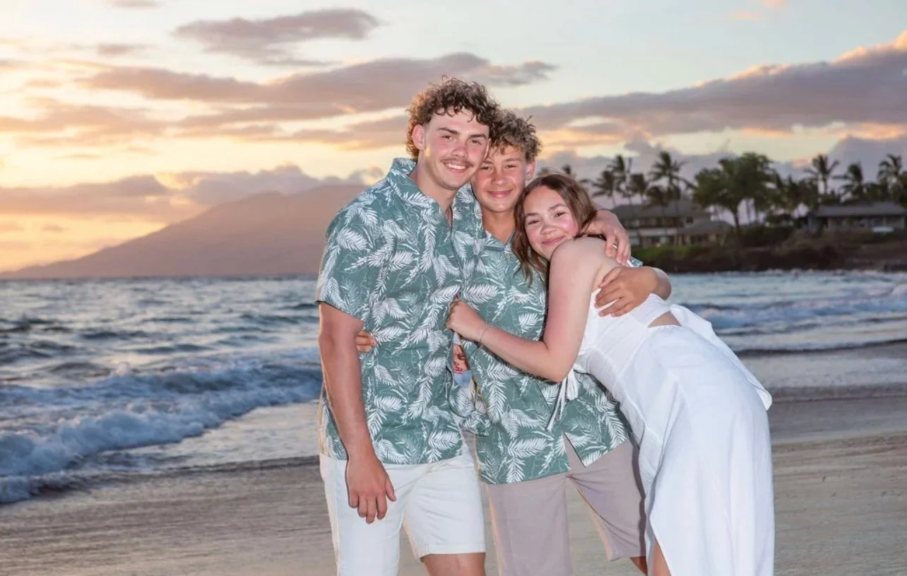 Three young friends hugging and smiling on the beach during sunset, with ocean waves and palm trees in the background.