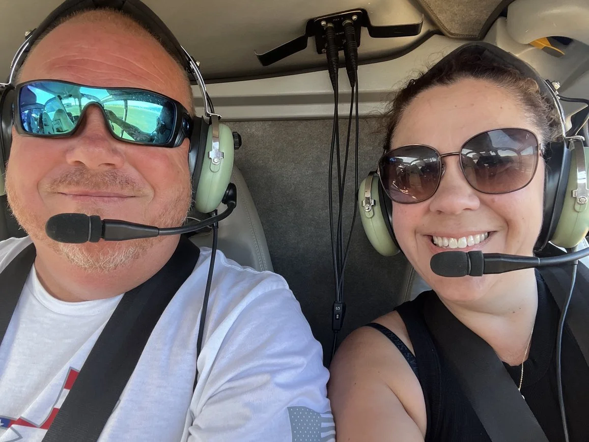 A man and woman inside a helicopter cockpit wearing headsets with microphones, sunglasses, and smiling at the camera.