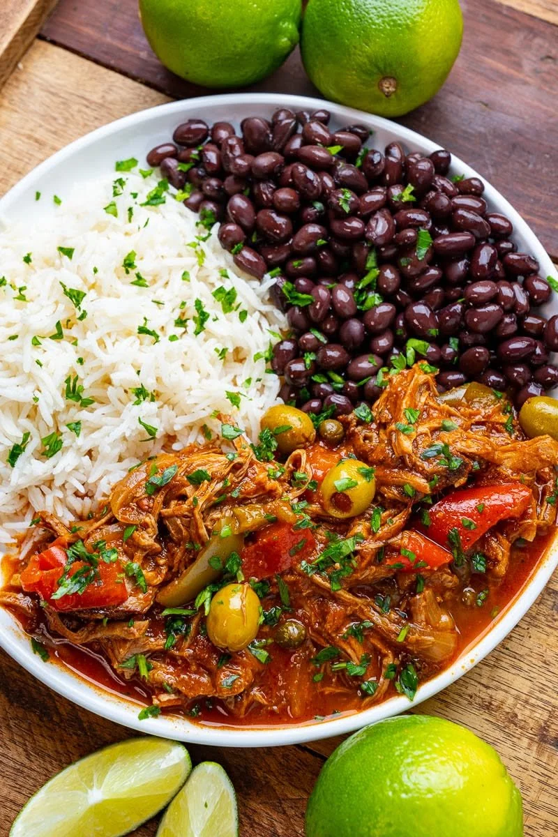 A white bowl filled with white rice, black beans, and a shredded meat stew with vegetables and herbs, placed on a wooden surface with lime wedges and whole limes nearby.