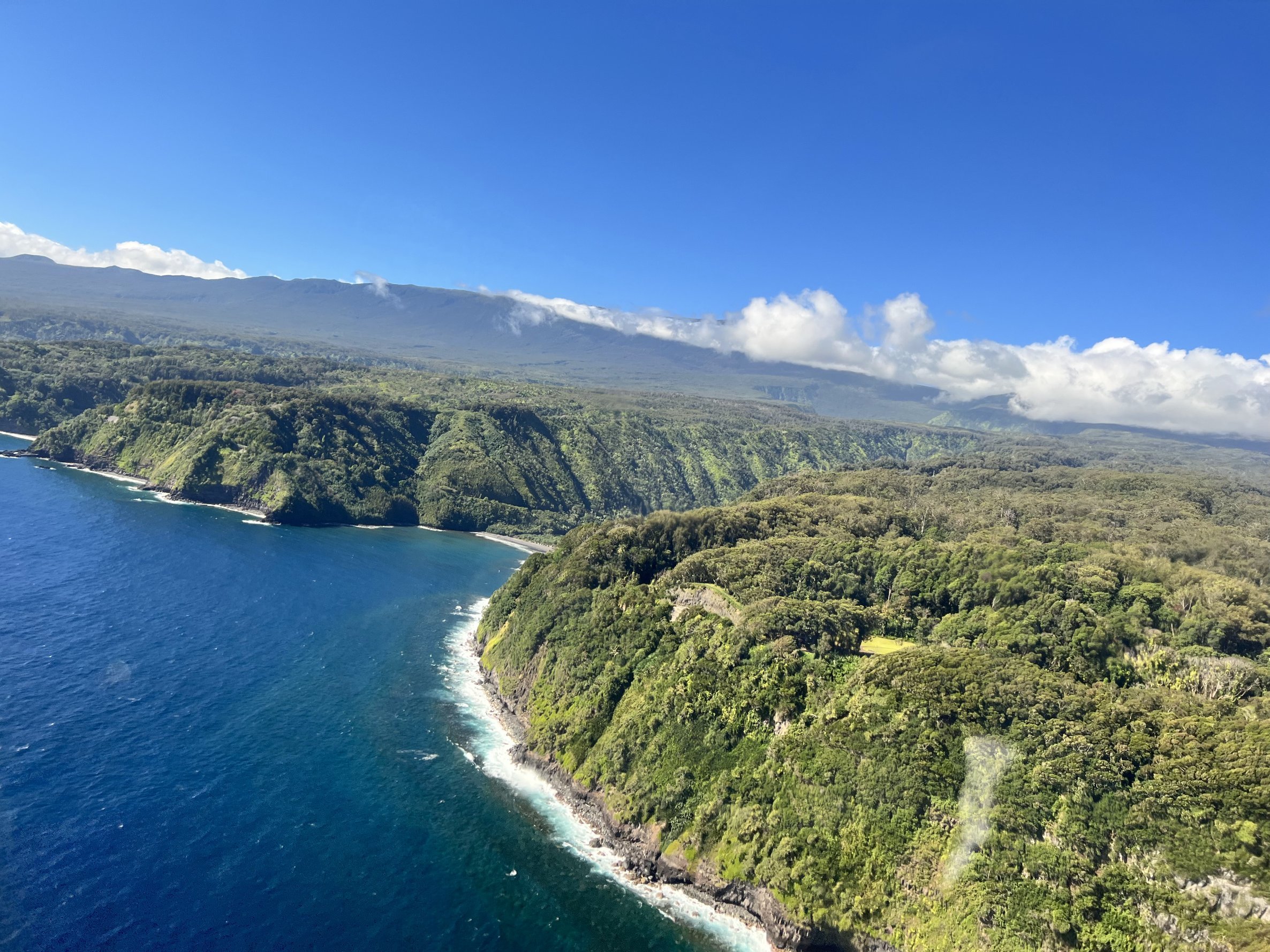 A scenic coastal landscape with lush green hills descending into the blue ocean, a clear blue sky, and clouds over a mountain in the background.