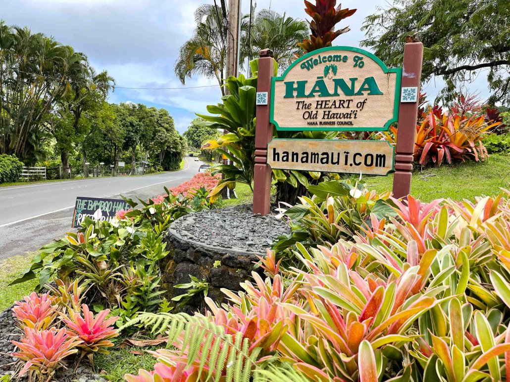 Welcome sign for Hana, Hawaii, with lush tropical plants and a road in the background.