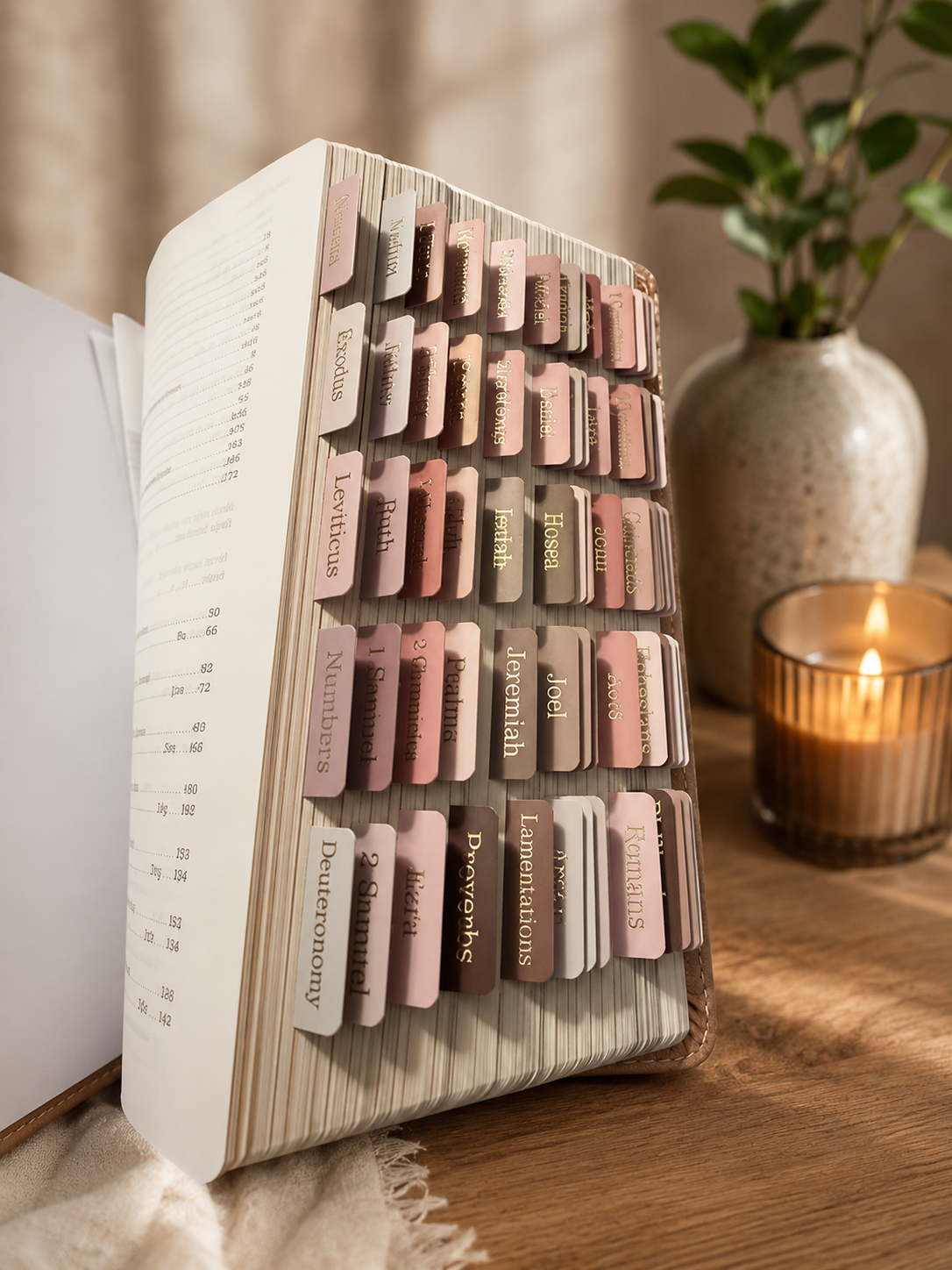 A Bible with colored tabs marking various books and sections, placed on a wooden surface beside a lit candle in a glass holder and a ceramic vase with green leaves.