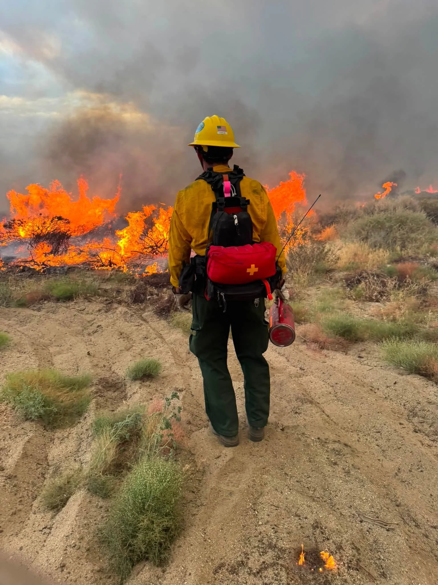 A firefighter wearing a yellow helmet and jacket, carrying a red backpack and a fire extinguisher, standing in front of a wildfire with orange flames and smoke on a dry, open landscape.