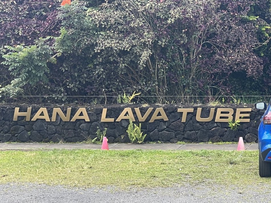 Sign that reads "HANA LAVA TUBE" in front of a garden wall with foliage and plants.