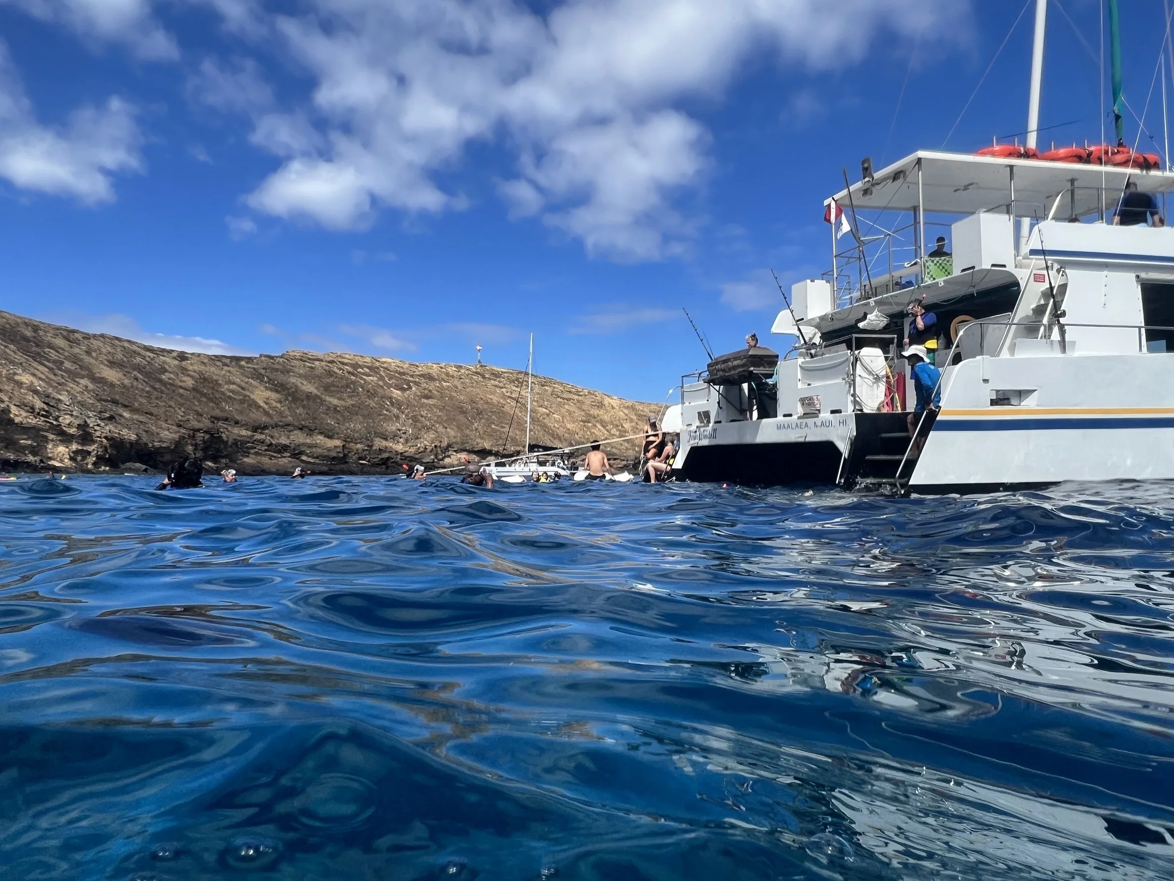 People swimming near a white boat anchored in the ocean close to a rocky hill under a blue sky with scattered clouds.