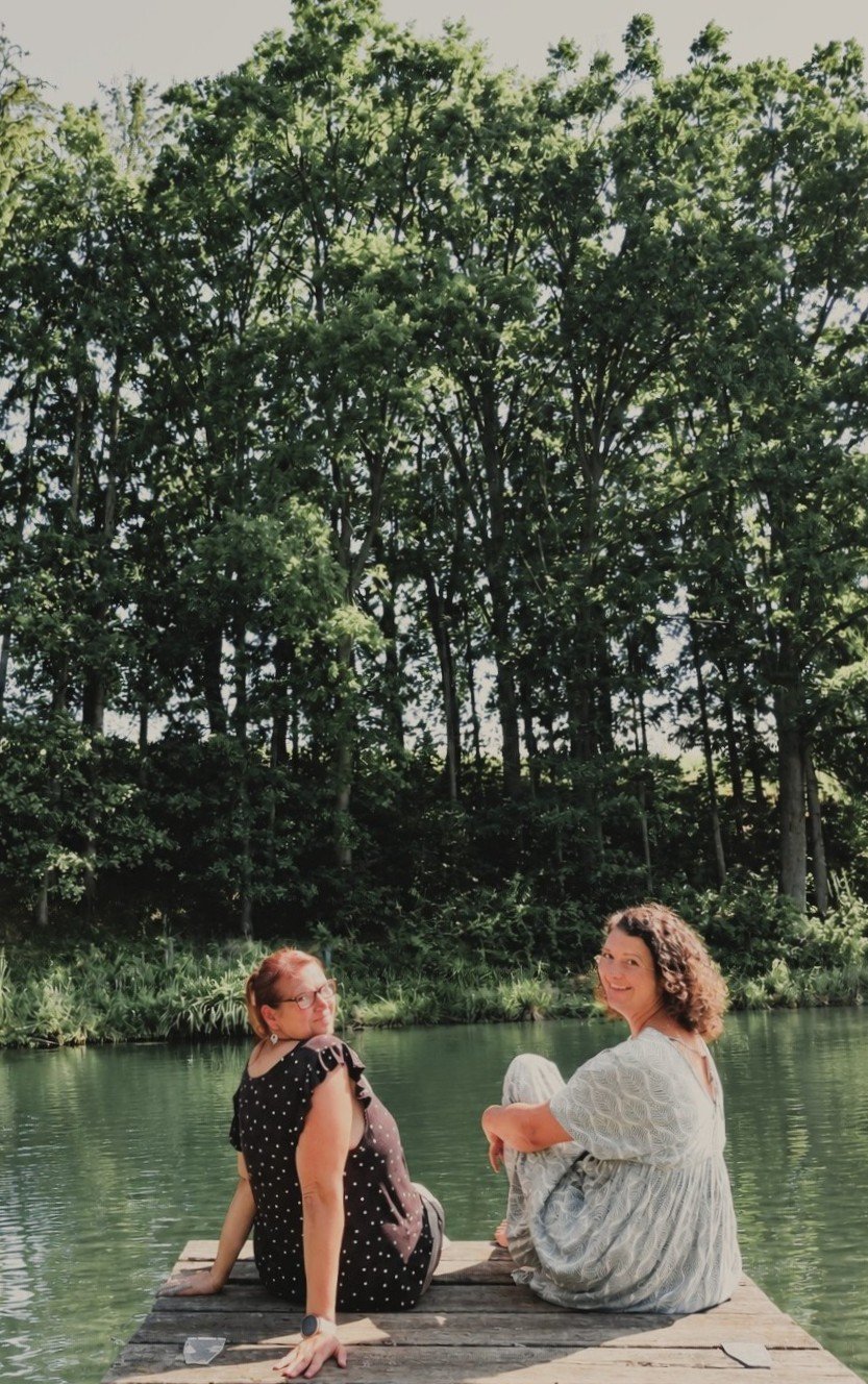 Zwei Frauen sitzen entspannt auf einem kleinen Holzsteg am Fluss, umgeben von grünen Bäumen, sonnenbeschienen und lachen.
