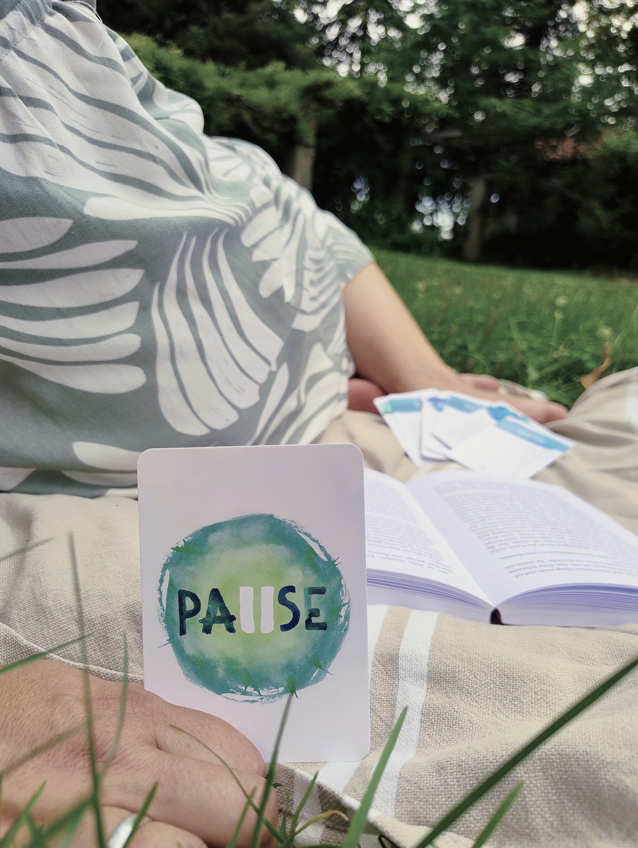 Ein Mensch sitzt im Freien auf einer Decke, liest ein Buch und hält eine Karte mit dem Wort 'PAUSE' und einem grünen, künstlerischen Kreis. Im Hintergrund sind Bäume und grünes Laub, es sieht nach Natur aus.