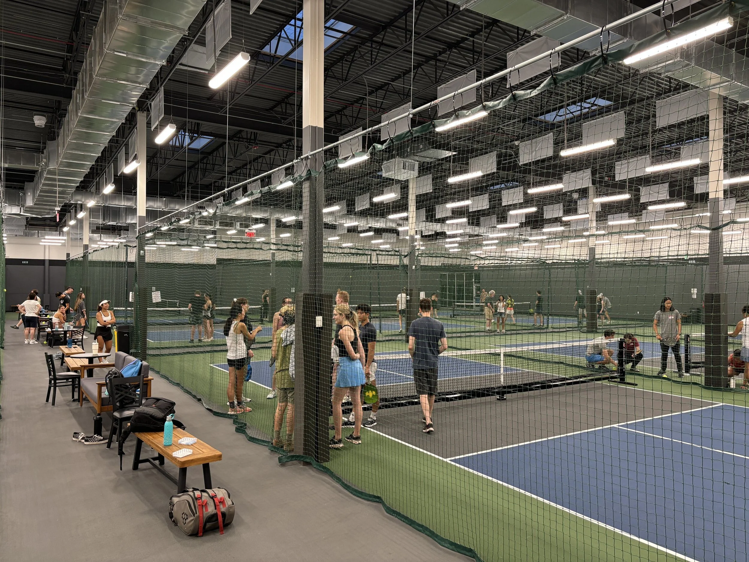 3rd Shot Pickleball Indoor pickleball courts with players and spectators, surrounded by a tall net, with benches and backpacks in the foreground.