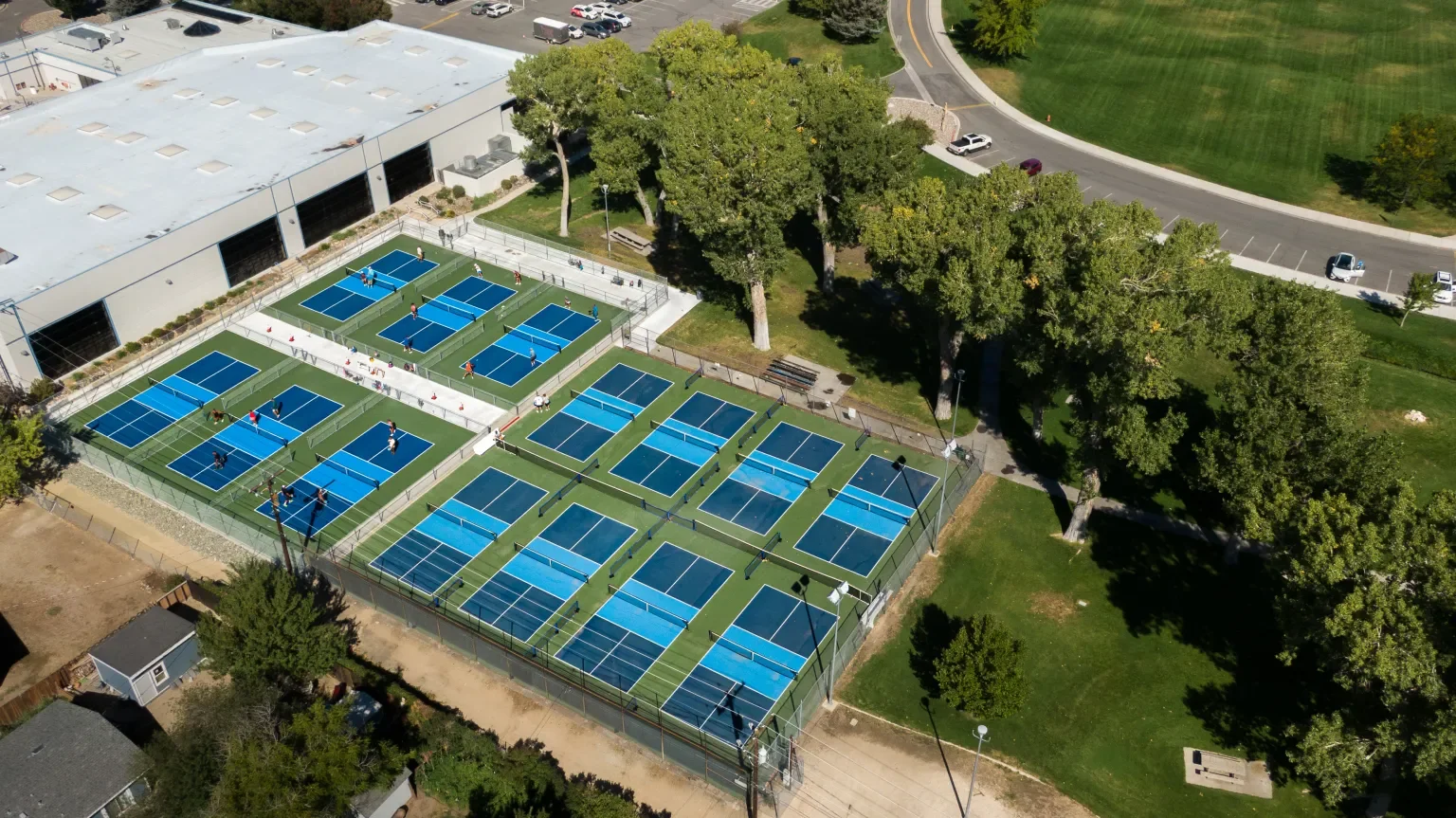 An aerial view of Mills Park pickleball courts with players, surrounded by trees and a grassy park area, adjacent to a large building with a parking lot.