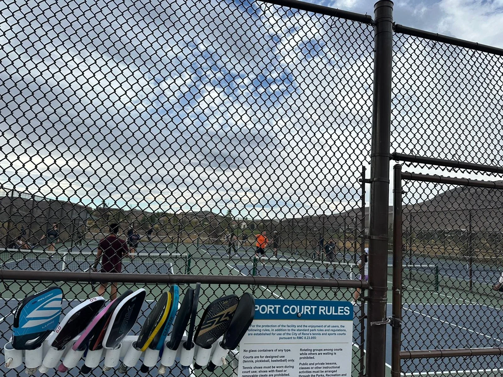 A view of Cyan pickleball courts with players playing a game, seen through a chain-link fence, with a row of pickleball paddles left in the paddle rack.