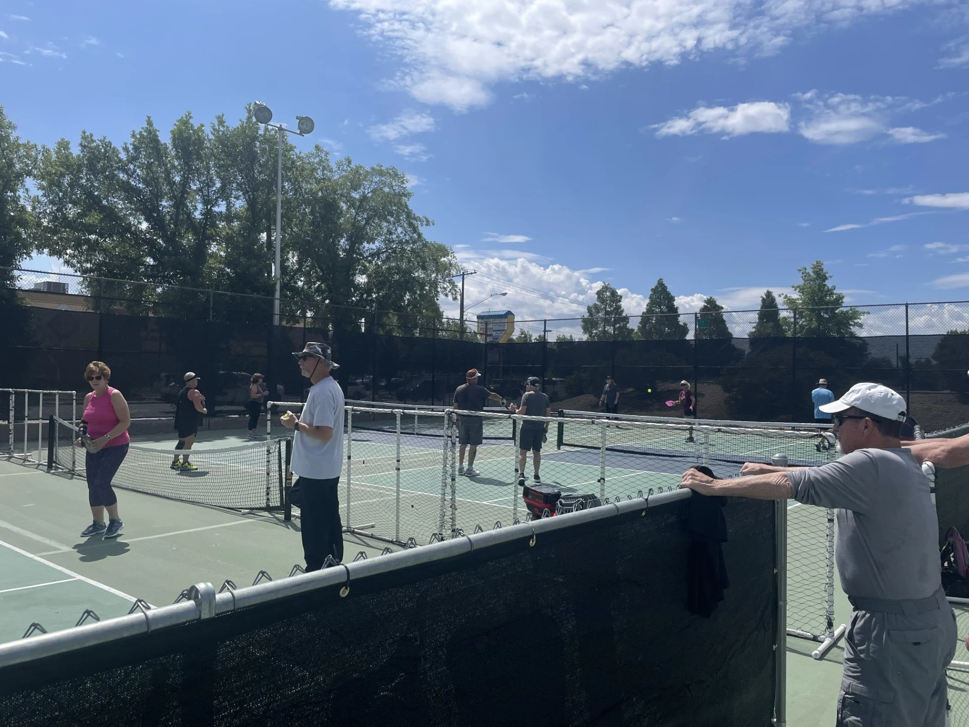 A group of people playing pickleball at Burgess Park pickleball courts under a partly cloudy sky. Some individuals are actively engaged in the game, while others are watching.