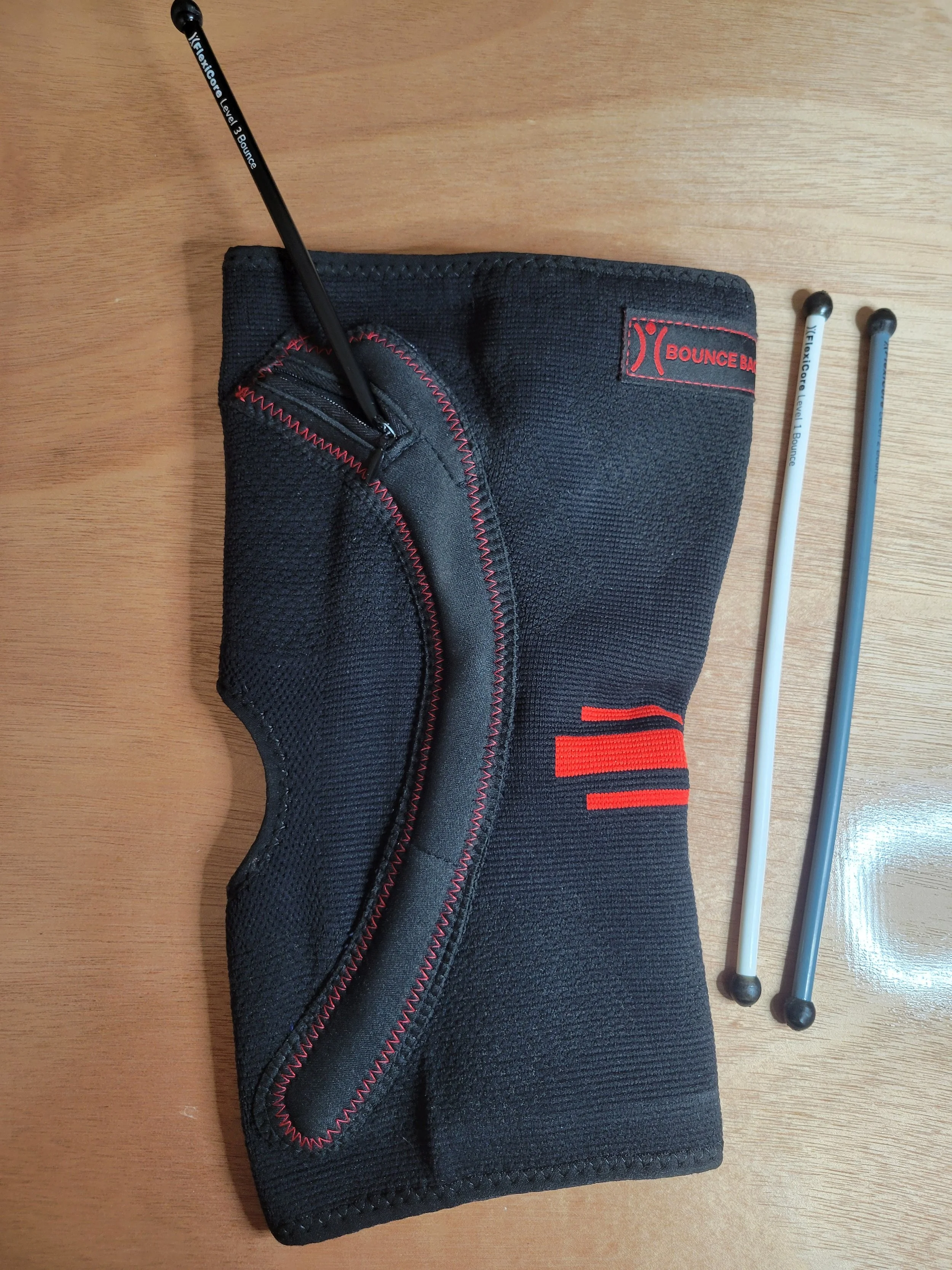 A black foam roller with red stitching, a zippered compartment, and the brand name 'Bounce Back' on a patch, placed on a wooden surface. Two massage sticks with black handles and rubber tips are beside it.