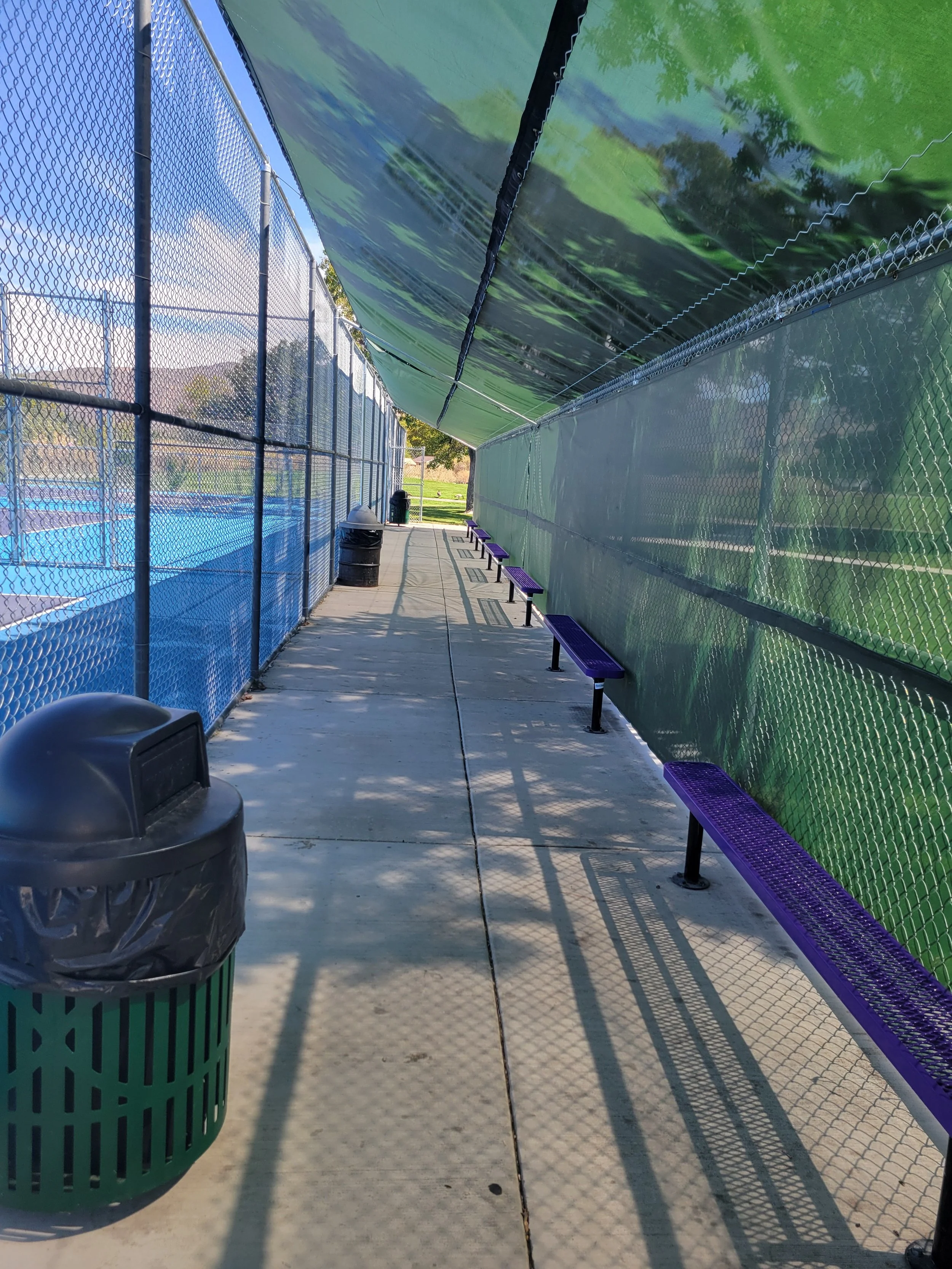 An outdoor shaded walkway area alongside the Mira Loma pickleball courts with chain-link fences on both sides, purple benches along the path, a green trash can, and large green shade cloths overhead casting shadows on the sidewalk.
