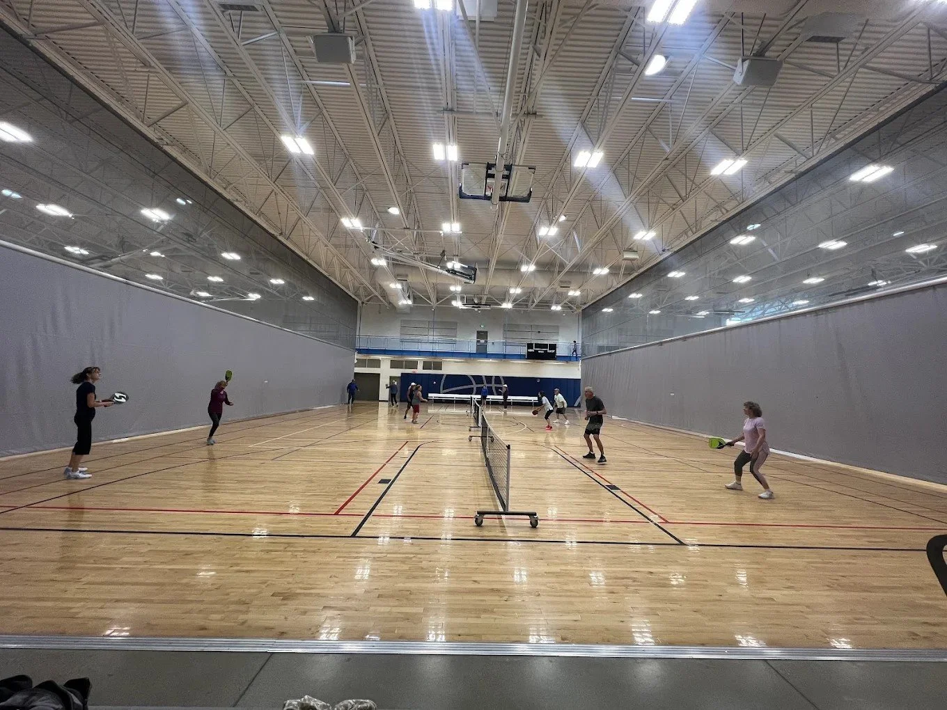People playing pickleball at the MAC indoor gym with a wooden floor, gray walls, and a high ceiling with bright lights.