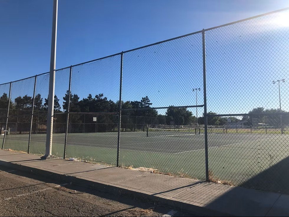 Oppio park pickleball courts enclosed by a chain-link fence under a clear blue sky, with trees and light poles in the background.