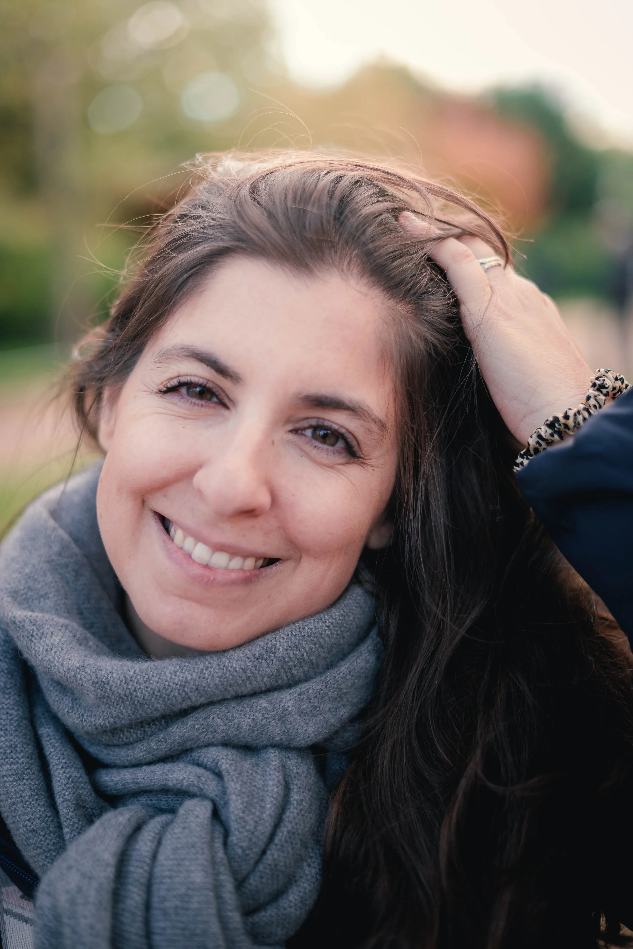 Close-up of a smiling woman with brown hair, wearing a gray scarf, outdoors with blurred greenery and soft sunlight in the background.