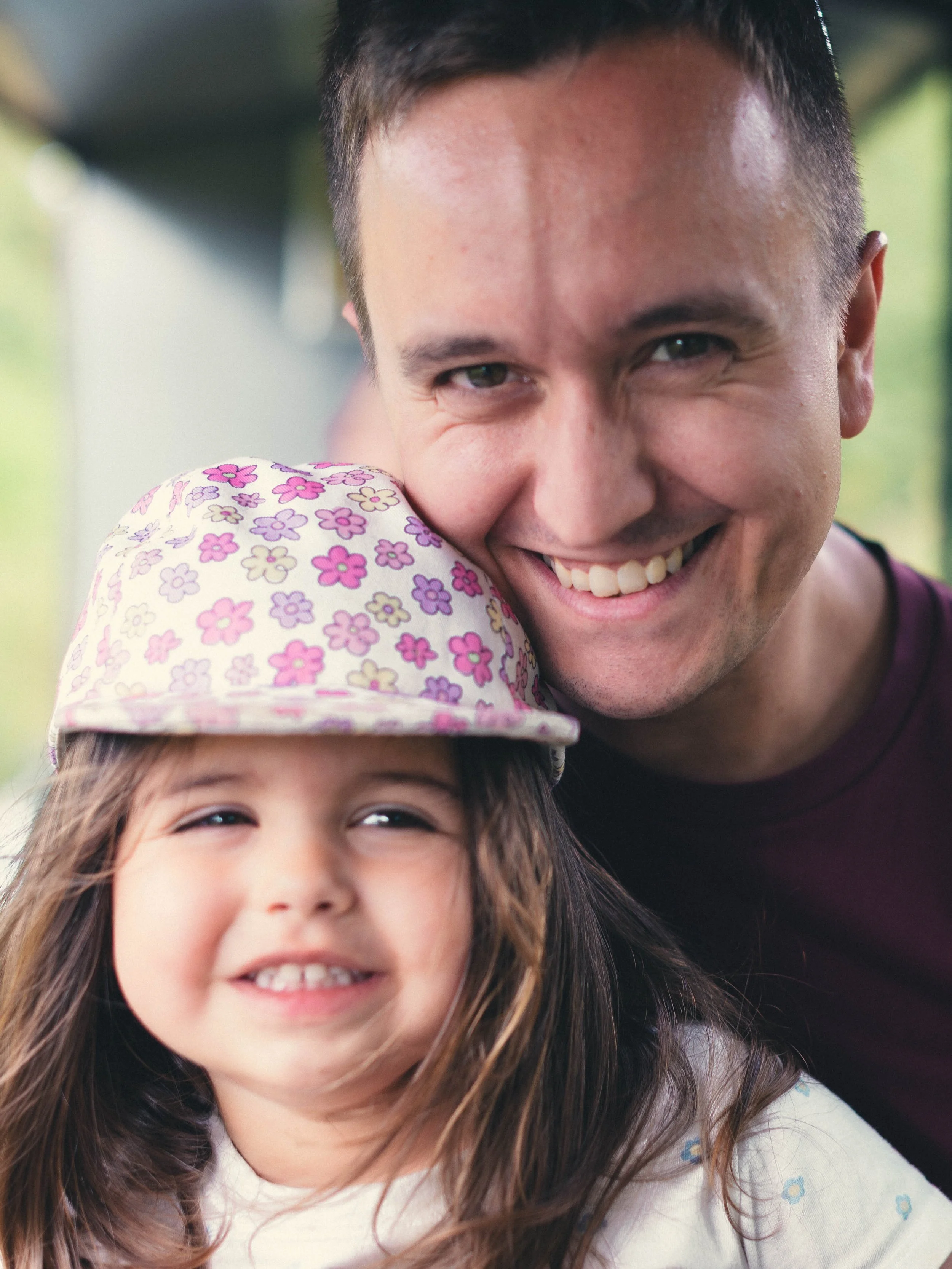 A young girl with long brown hair wearing a pink polka dot bucket hat and an off-white shirt with small blue flowers, smiling at the camera, with a man smiling in the background.