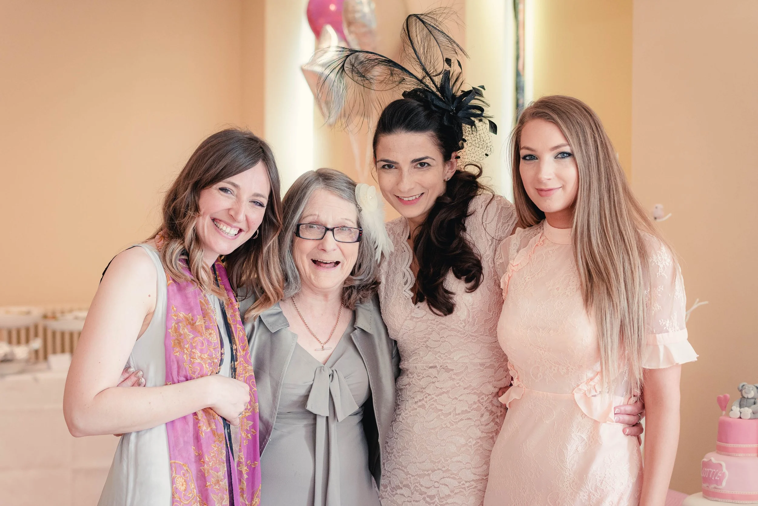 Four women smiling in a group, celebrating a baby shower, with a pink and pastel themed background.