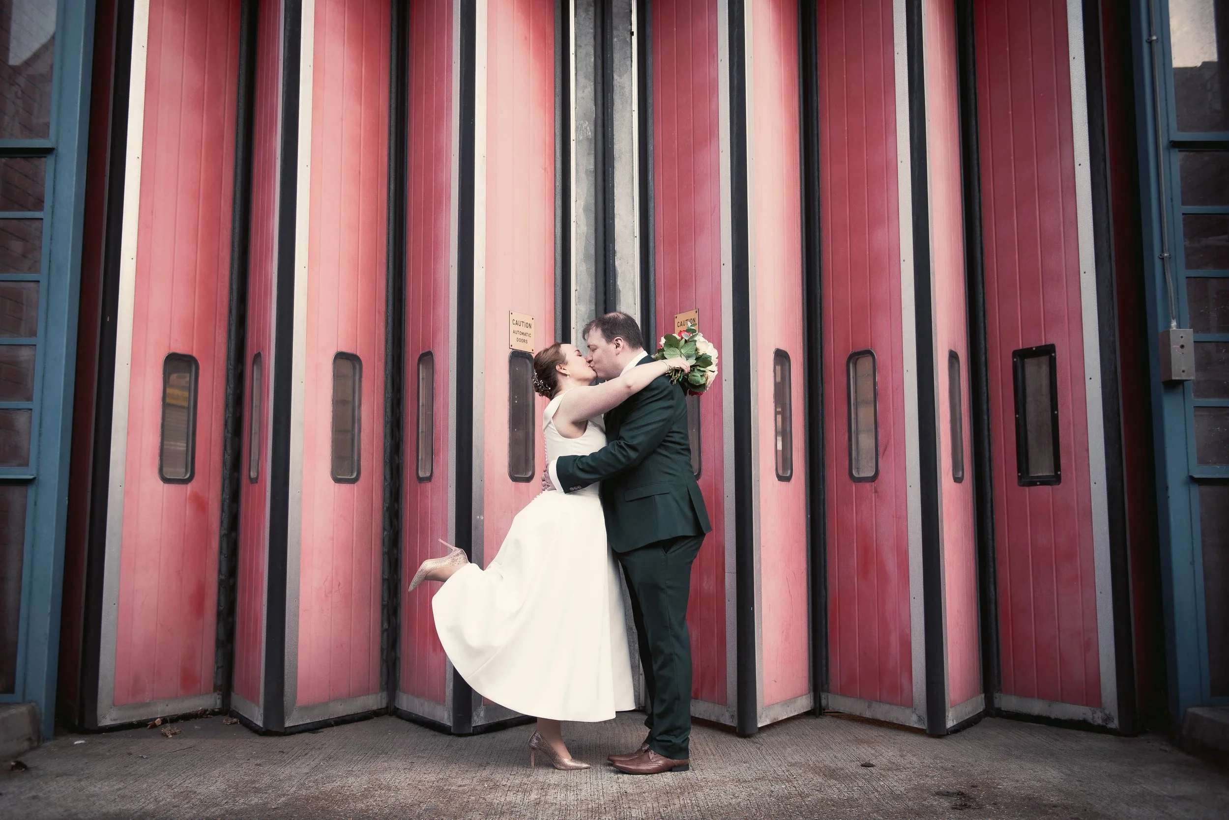A newlywed couple sharing a kiss in front of a red and gray industrial door, with the bride holding a bouquet of flowers and both dressed in wedding attire.