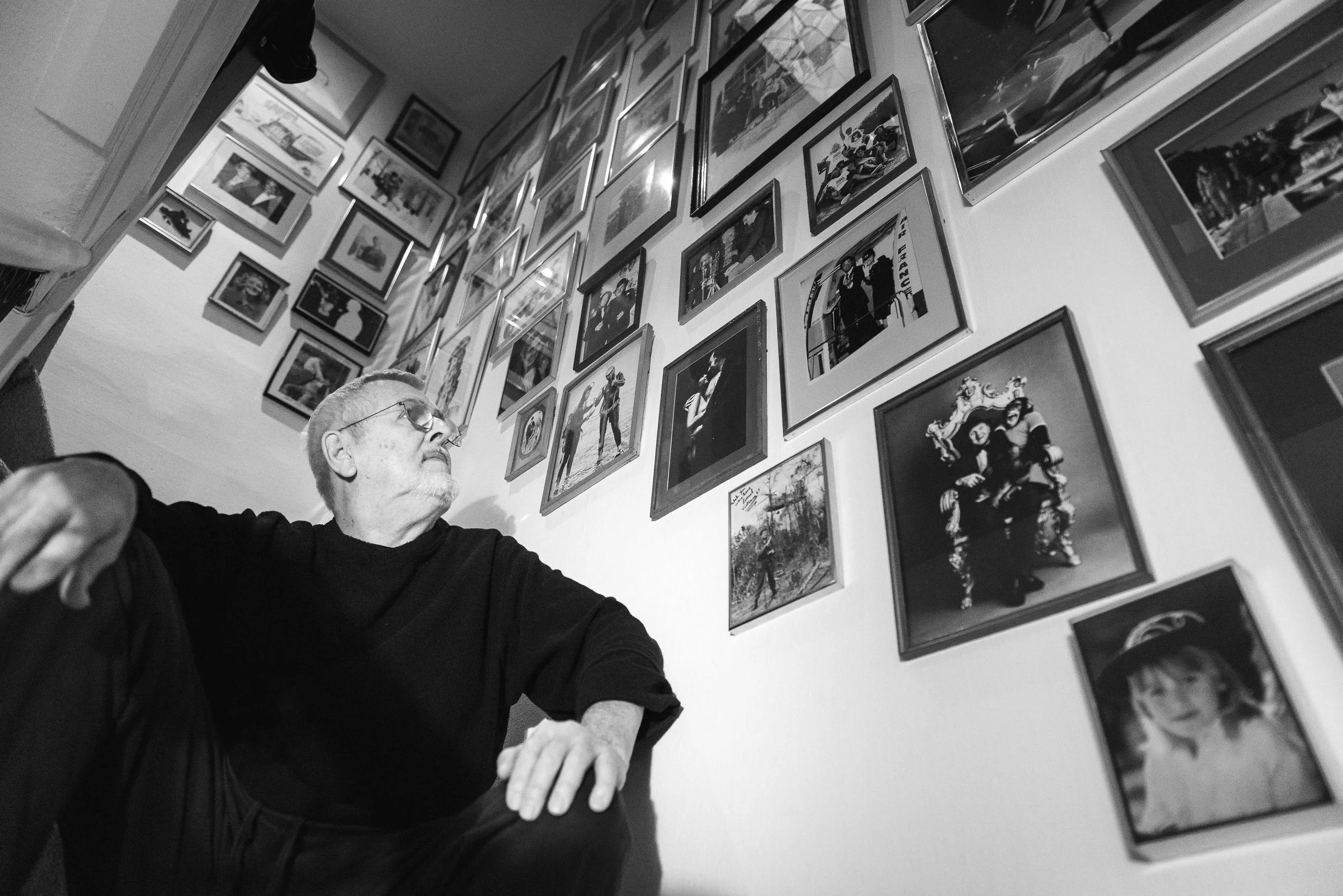 Black and white photo of an elderly man with glasses sitting at a table, with a gallery wall of framed photographs behind him, some of which show people and events.