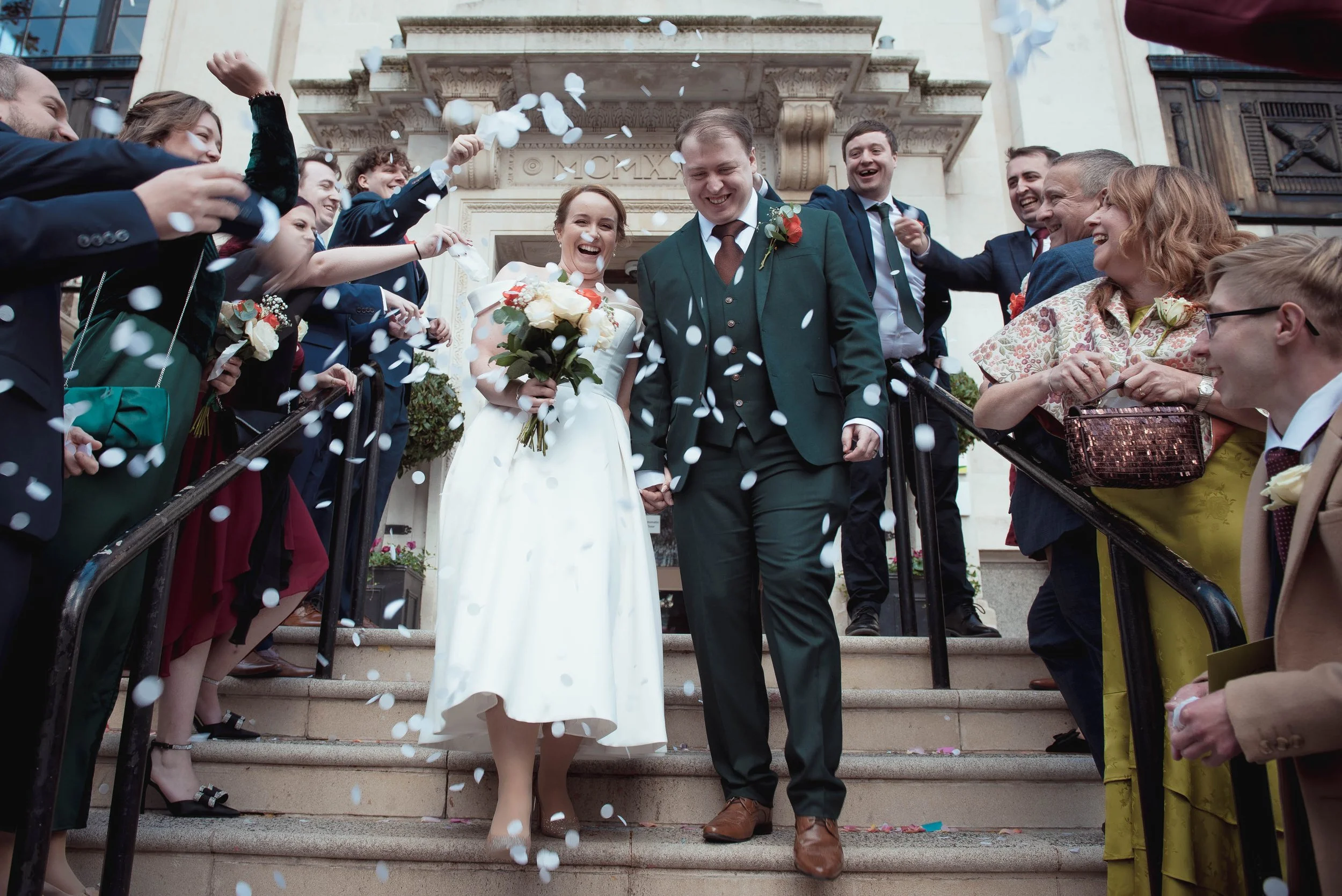 A bride and groom walking down the stairs after their wedding ceremony, surrounded by friends and family throwing white confetti, outside a building with classical architectural details.