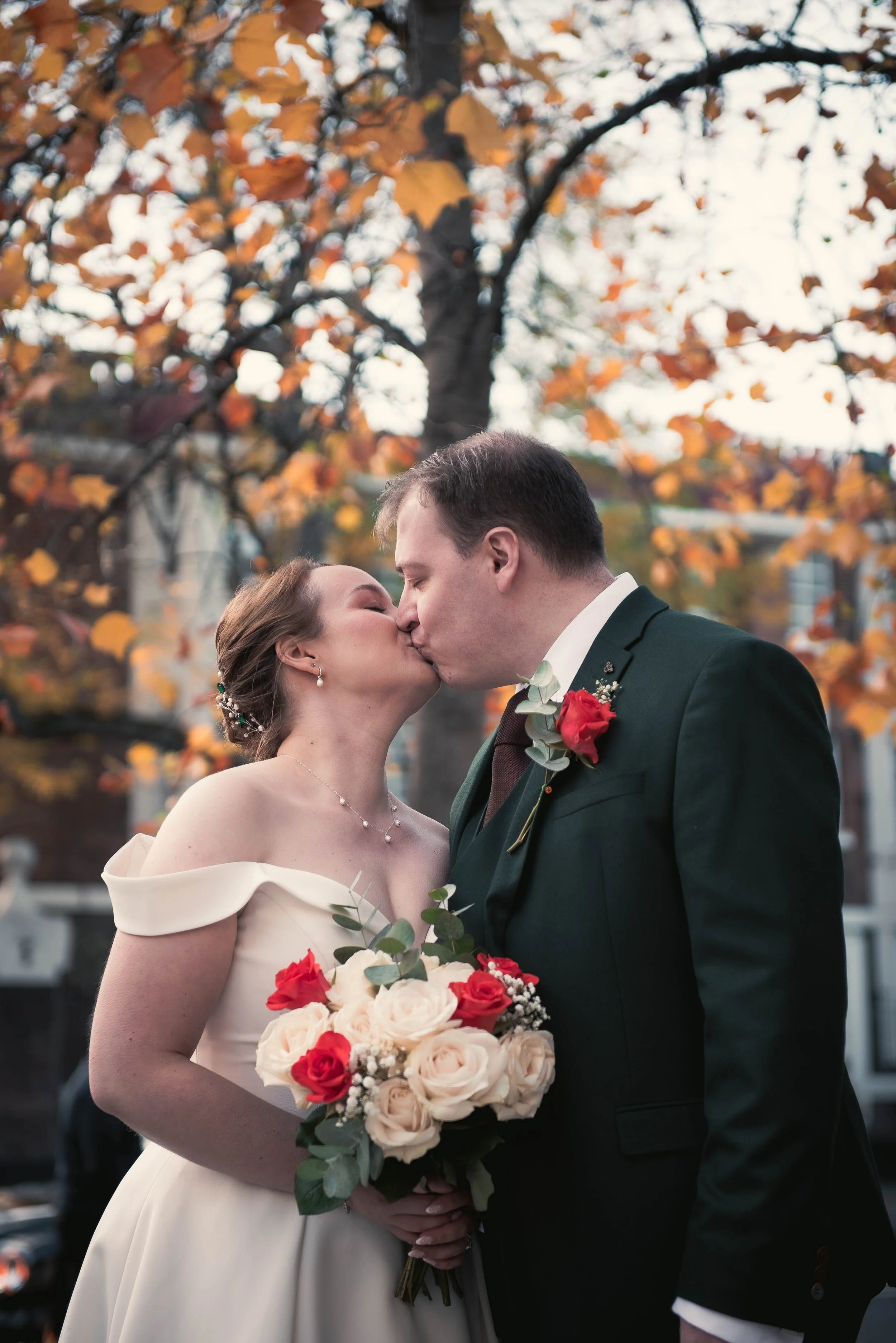 A bride and groom sharing a kiss outdoors during their wedding, with autumn trees and orange leaves in the background. The bride is holding a bouquet of cream and red roses.