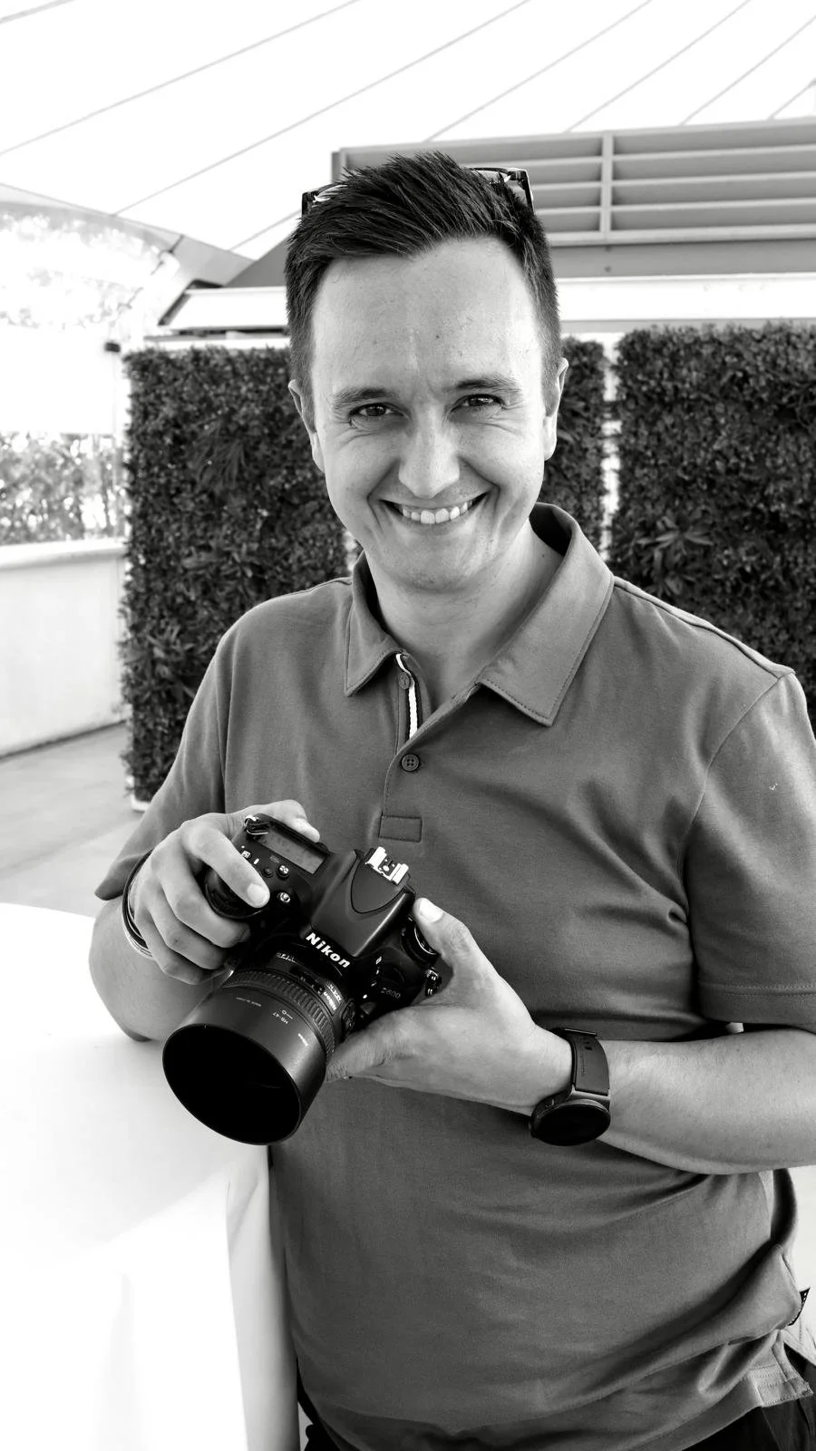 A smiling man holding a Nikon camera outdoors with a hedge and part of a modern building in the background.