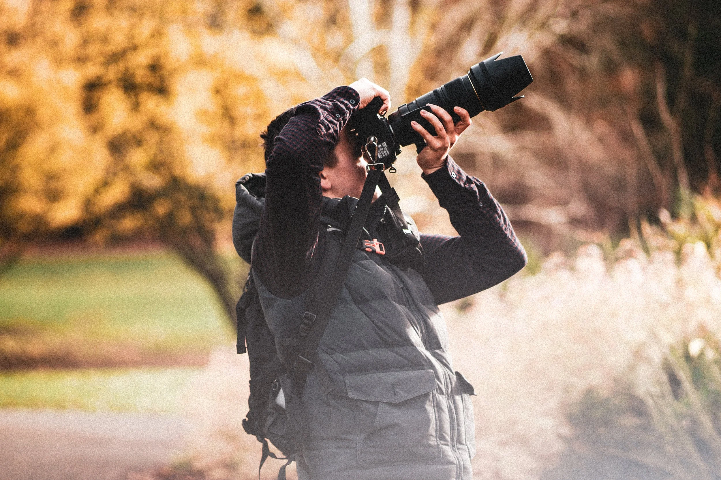 A person in outdoor clothing is taking a photograph with a camera that has a large telephoto lens, outdoors with autumn-colored trees in the background.