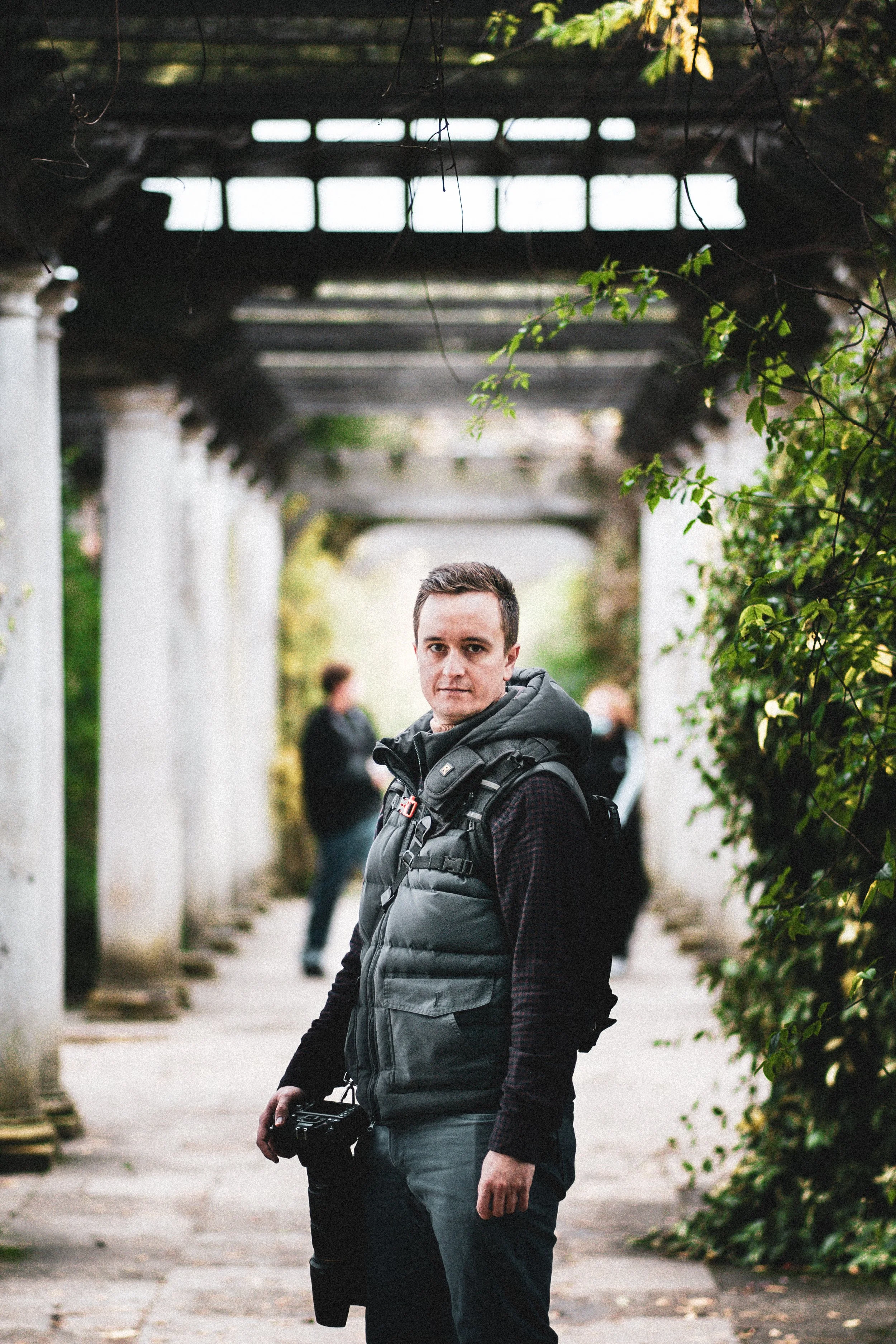 A man wearing a gray vest and black long-sleeve shirt, holding a camera, stands outdoors under a wooden pergola with green foliage on the side. Other people are blurred in the background.