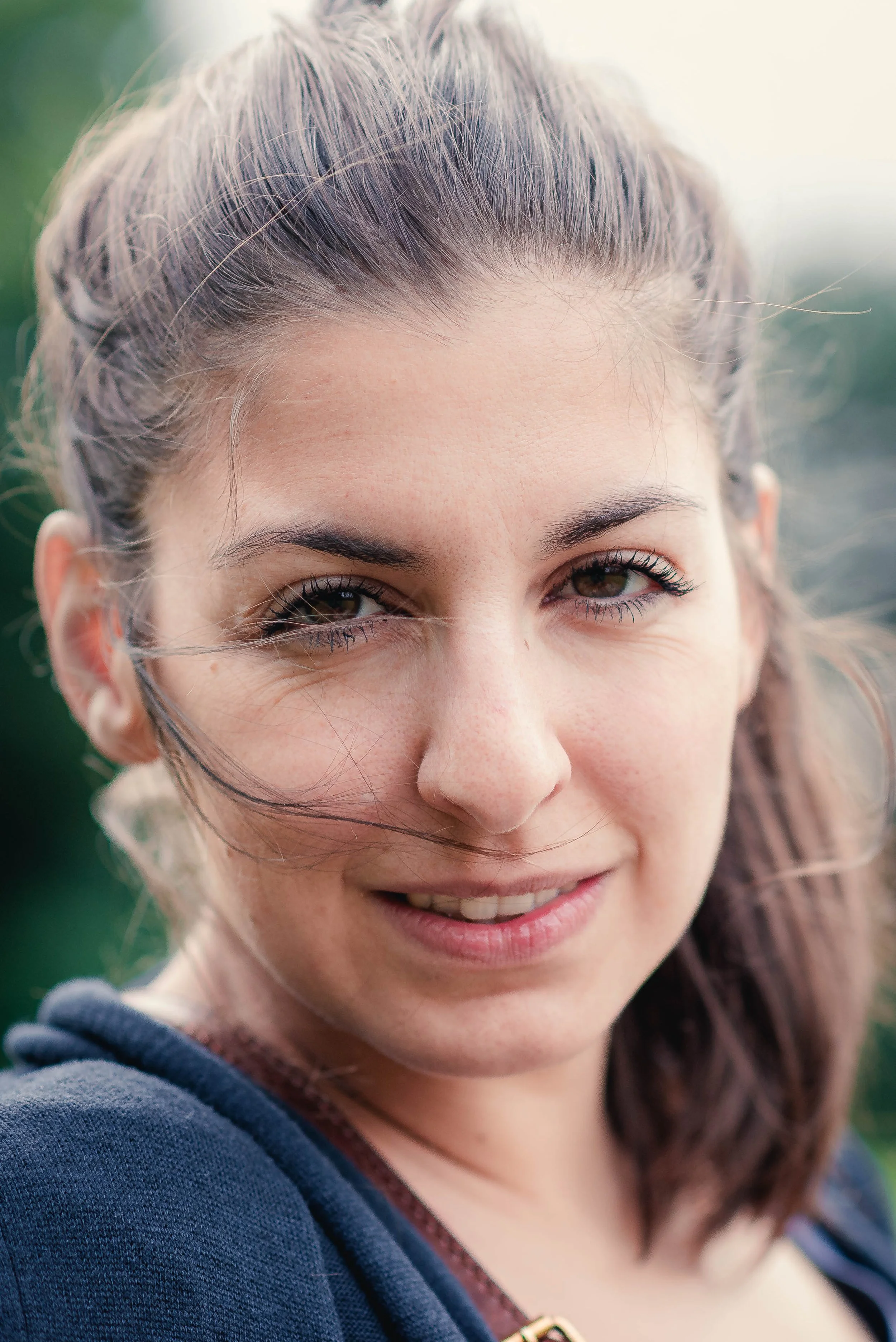 Close-up of a young woman smiling, with chestnut hair, wearing a navy blue hoodie, outdoors with blurred greenery background.