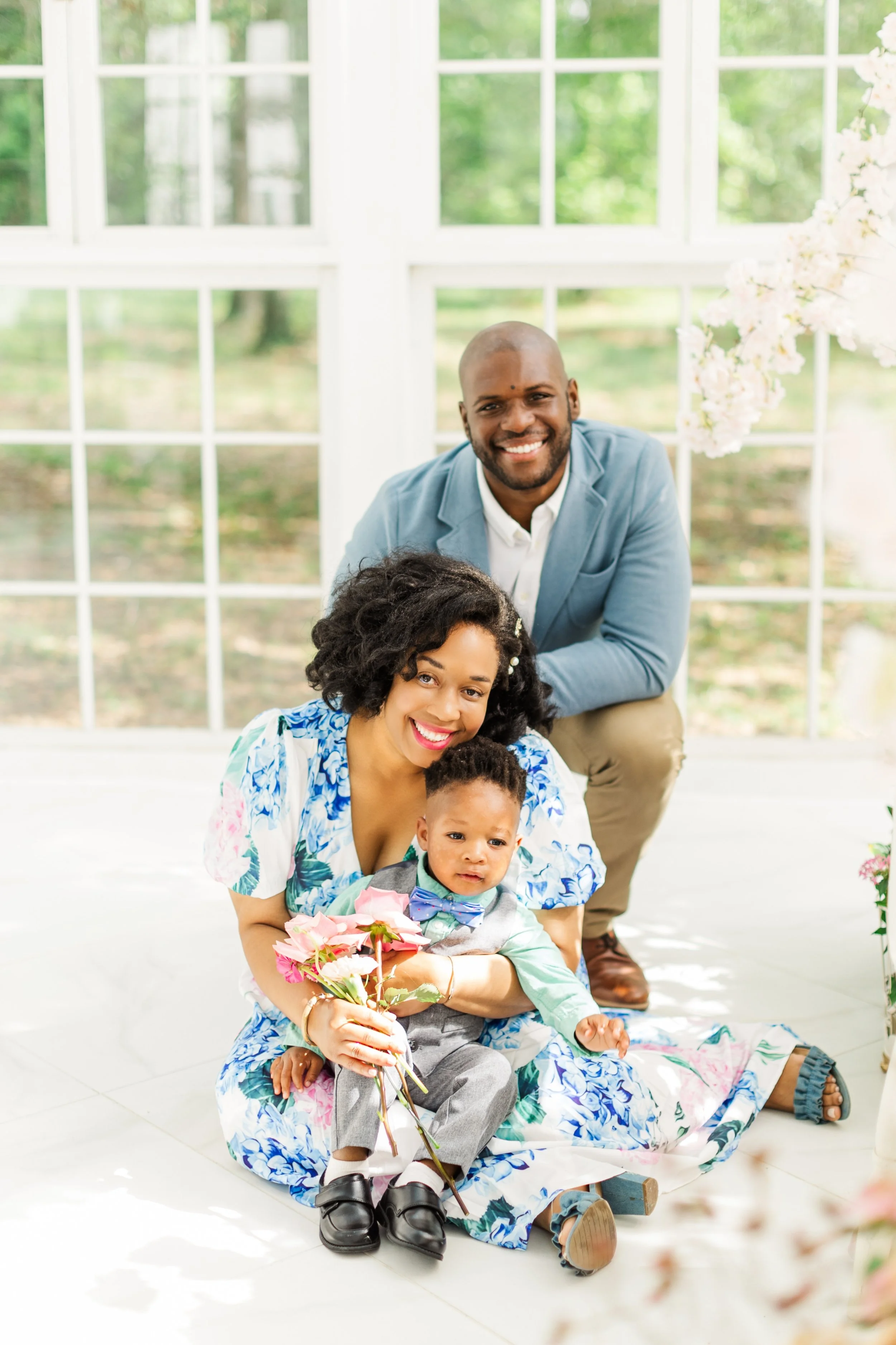 Family group photos in a bright room with large windows and green outdoor scenery, including a smiling man, a woman holding a child, and a young boy with flowers. The woman is in a patterned dress, and the man is in a blue blazer.