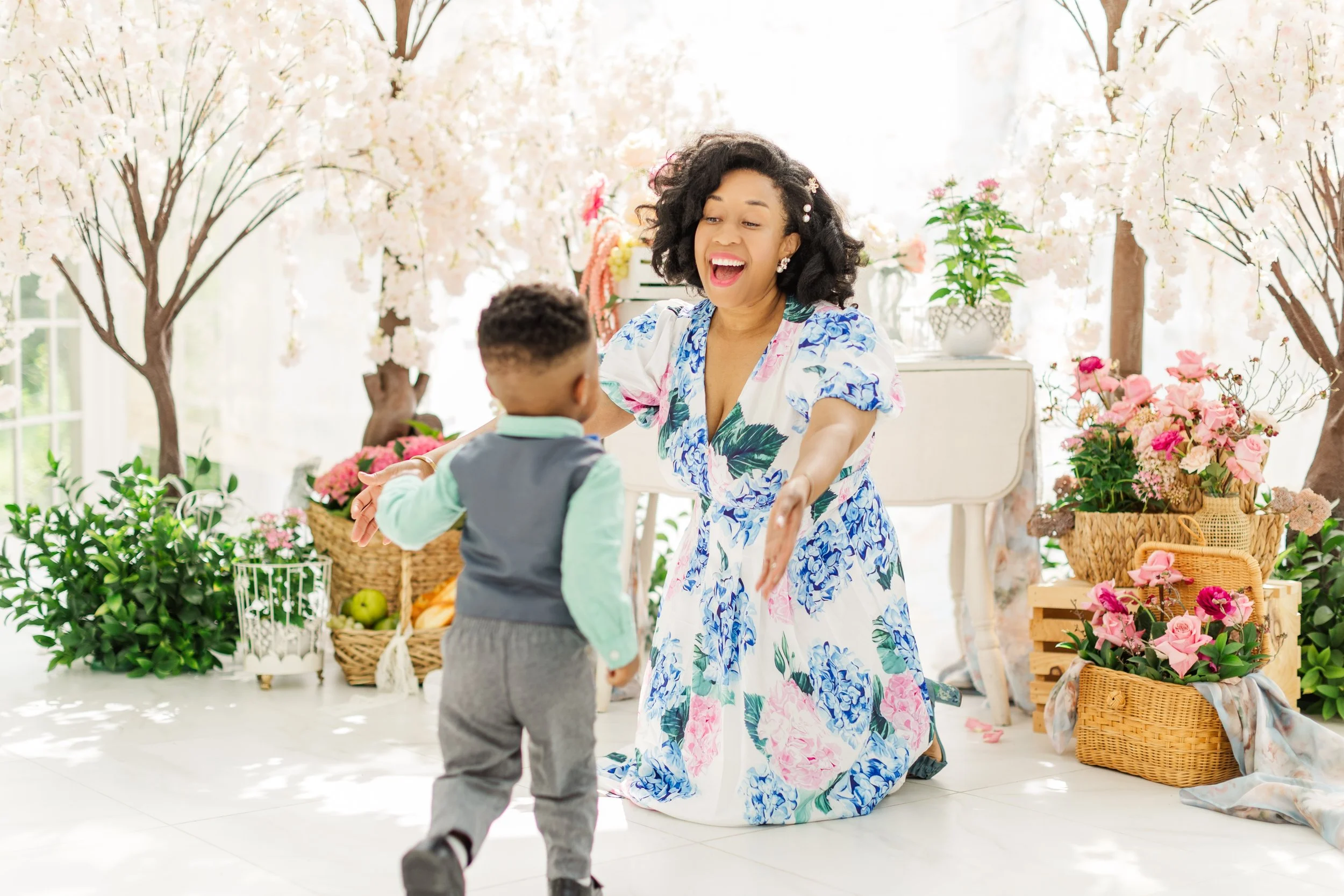 A woman dancing happily with a child in a flower shop filled with pink and white blossoms and baskets of flowers.