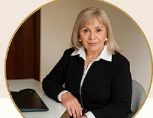 Mujer de cabello rubio y peinado corto, vestida con blazer negro y camisa blanca, sentada en escritorio con teléfono y papeles.