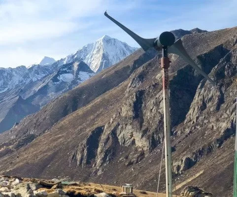 Viento en un paisaje montañoso con picos nevados al fondo, y un aerogenerador en primer plano.