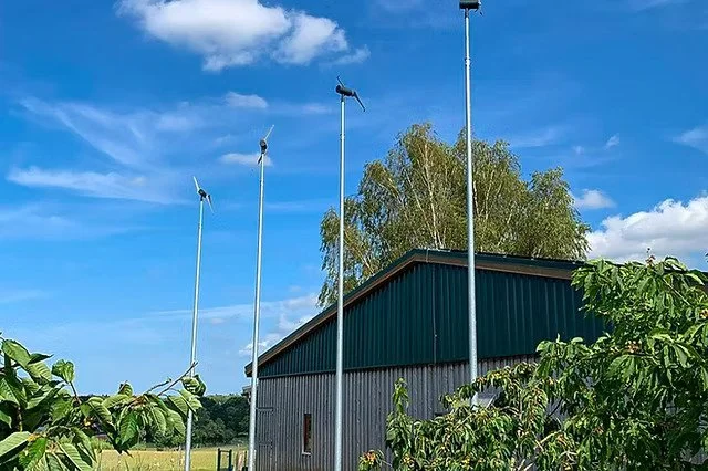 Cuatro molinos de viento en un campo junto a un edificio con paredes de metal y techos verdes, con árboles y arbustos alrededor, bajo un cielo azul con nubes.
