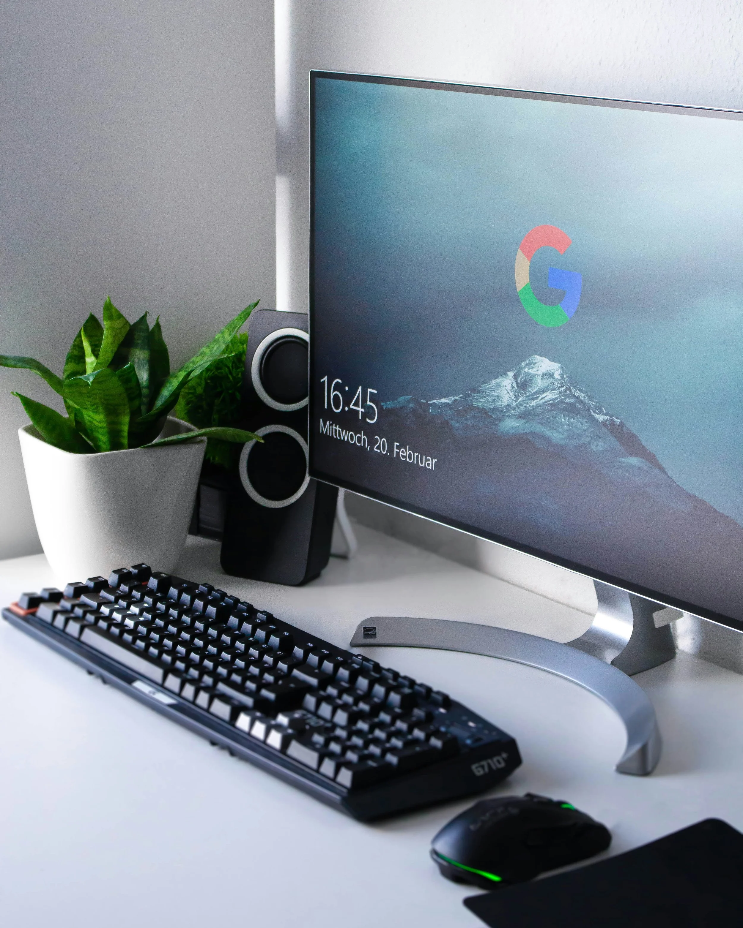 Computadora con pantalla grande, teclado, ratón con luce oscura y verde, planta en maceta blanca, y algunos objetos en un escritorio blanco.