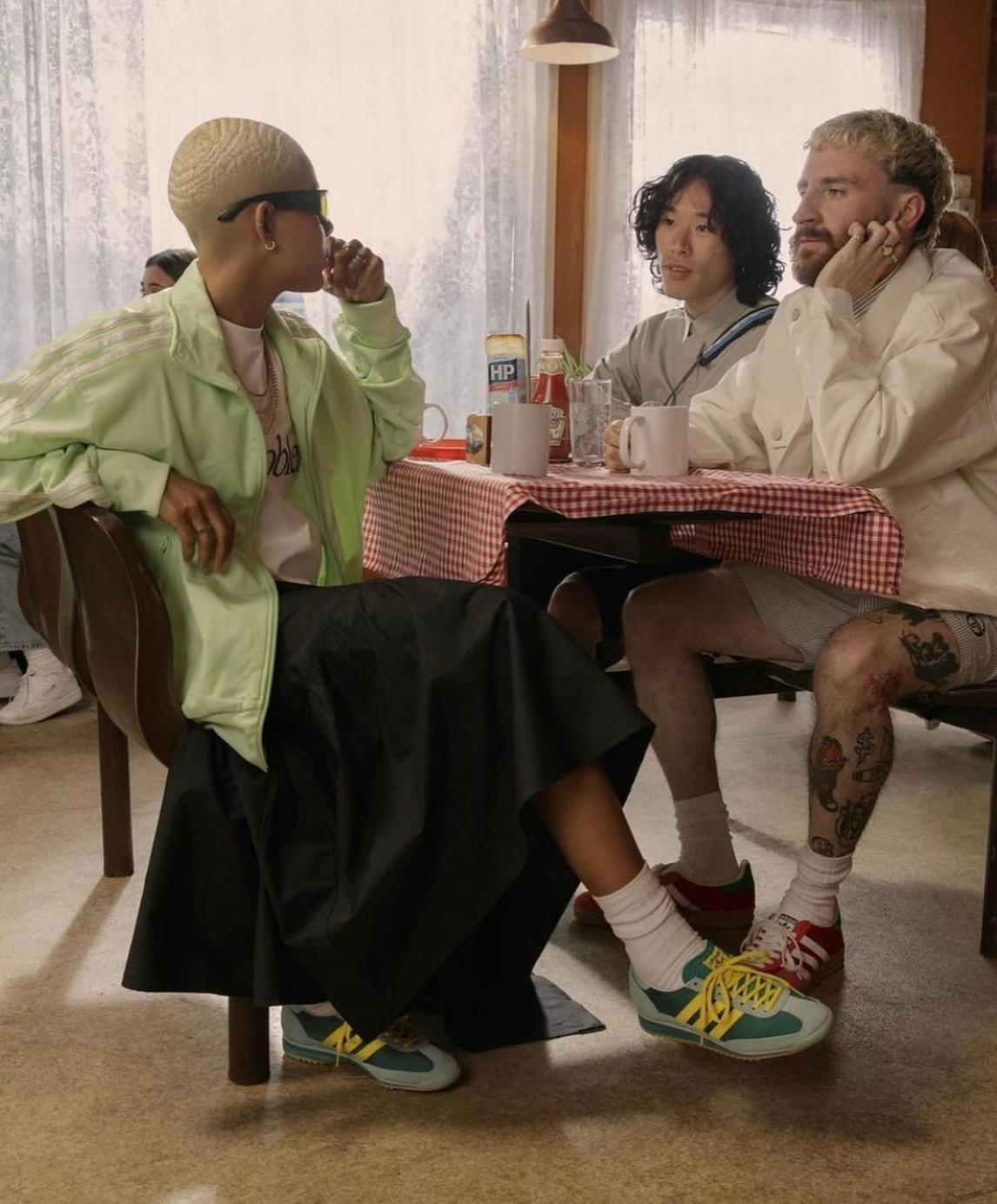 Three people sitting at a dining table in a casual restaurant. Two men and a woman are engaged in conversation, with various ketchup and mustard packets on the table. The woman on the left has a shaved head, sunglasses, and is wearing a light green jacket. The man on the right has tattoos on his legs, blonde hair, and is wearing a white jacket, shorts, and colorful sneakers.