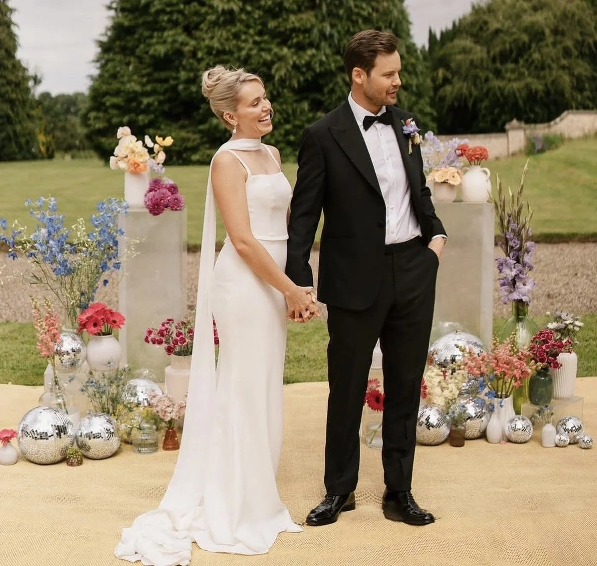 A bride and groom holding hands and smiling during their outdoor wedding ceremony, surrounded by colorful flowers and decorative mirrors.