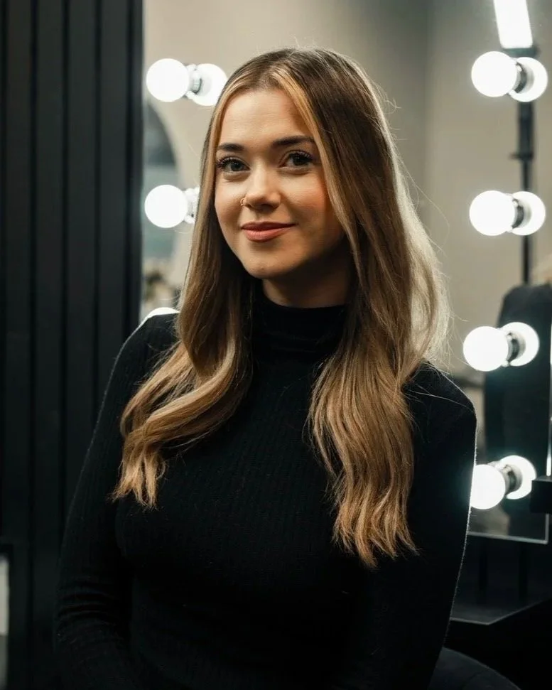 A young woman with long wavy hair, wearing a black turtleneck, standing in front of a mirror with bright vanity lights.